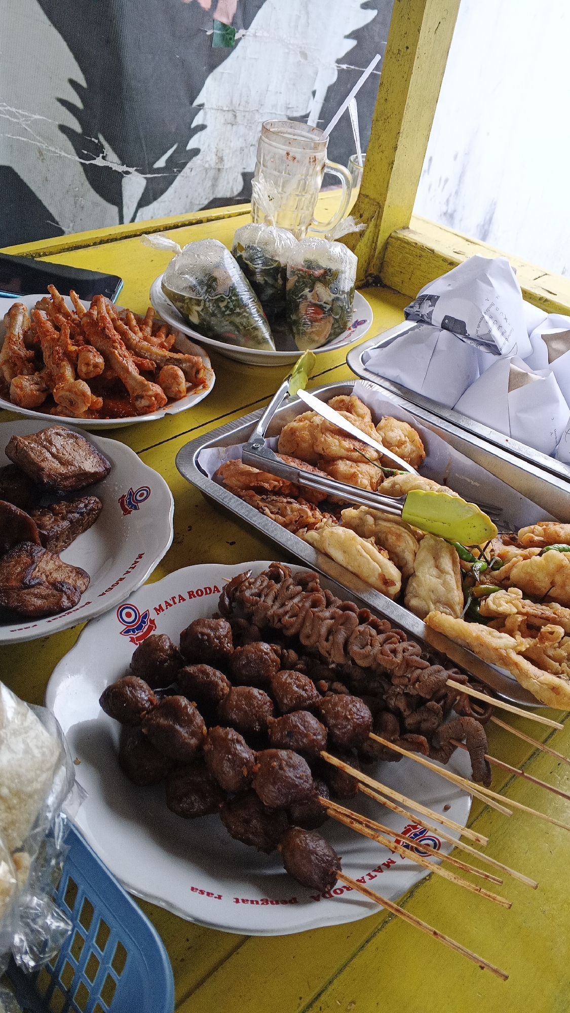 A close-up, top-down shot captures a vibrant street food spread on a bright yellow table, likely in Yogyakarta, Indonesia. The scene is dominated by a variety of Indonesian delicacies. In the foreground, a white plate displays skewers of dark, grilled meatballs and what appear to be intestines, both glistening with a savory sauce. To the left, another white plate holds several pieces of fried tofu and what looks like fried chicken wings coated in a reddish spice.
Further back, a tray overflows with an assortment of fried items, including battered pieces of fish or tofu, and some vegetables. Three small, clear plastic bags containing leafy green vegetables are placed on a smaller white plate. A partially empty glass with a straw sits behind these bags, suggesting a refreshing drink was consumed. The background features a textured wall with a graphic of a black bird or wing, partially obscured by a yellow wooden frame, hinting at an outdoor, possibly open-air, dining setting. The overall impression is one of a casual, delicious, and authentic Indonesian street food experience.