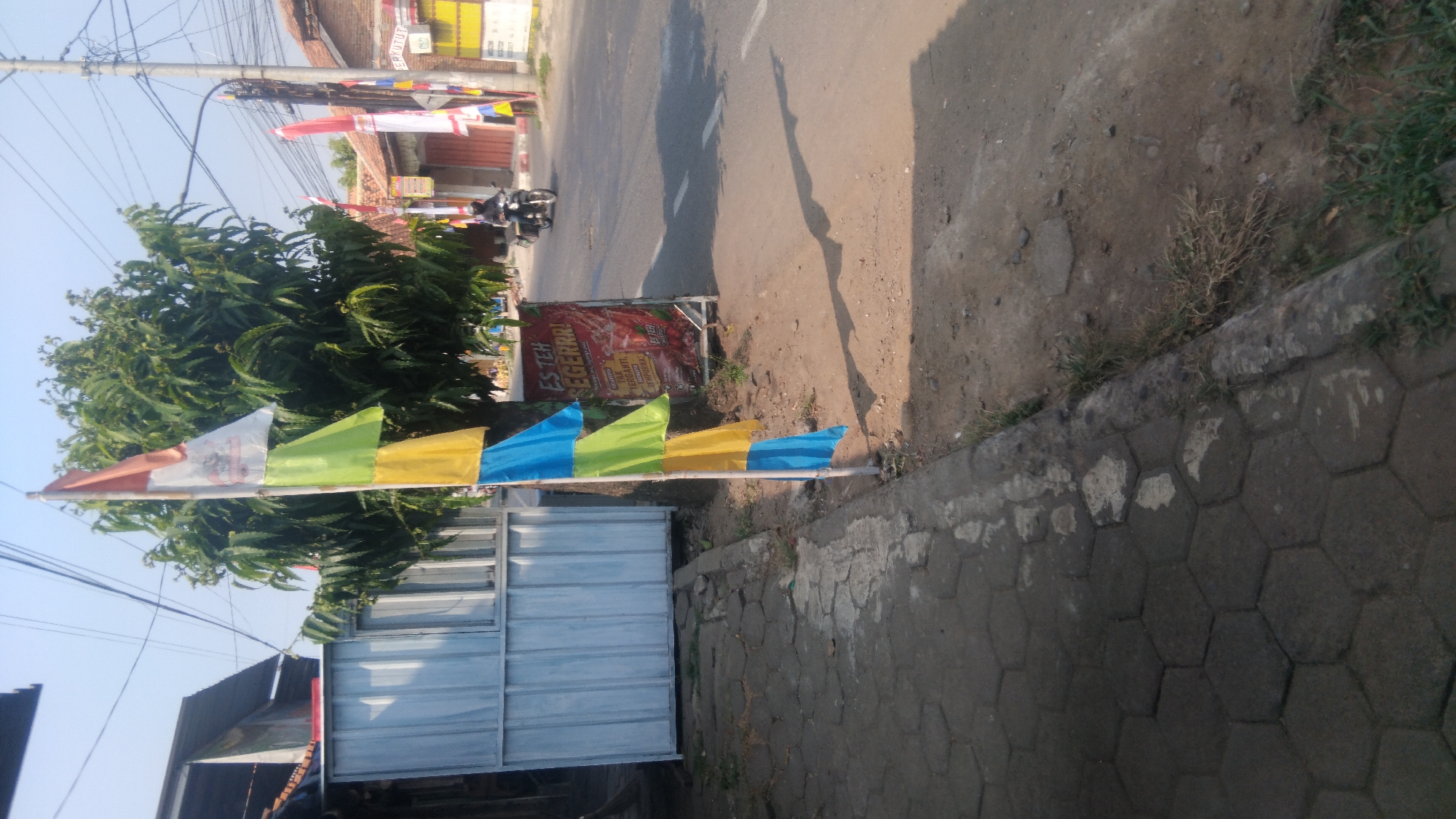 The image captures a street scene in Proyonanggan Tengah, Indonesia, under a clear sky. In the foreground, a tiled sidewalk runs along the left side of the image, with a tree and a flagpole with colorful banners planted in the ground beside it. The banners are layered and appear to be made of plastic or nylon, with colors including white, red, yellow, green, and blue. A small booth or structure with a white metal facade is visible behind the sidewalk.
Further back, a paved road stretches into the distance, with a motorcycle rider in motion. Several other flags, primarily red and white, are planted along the roadside. Commercial buildings and houses line the street, some with red tile roofs. A sign advertising "ES TEH SEGERRR!" (ice tea refresh!) is visible near the base of the tree, featuring an image of a beverage. The overall environment suggests a typical urban setting in Indonesia with everyday commercial activity.