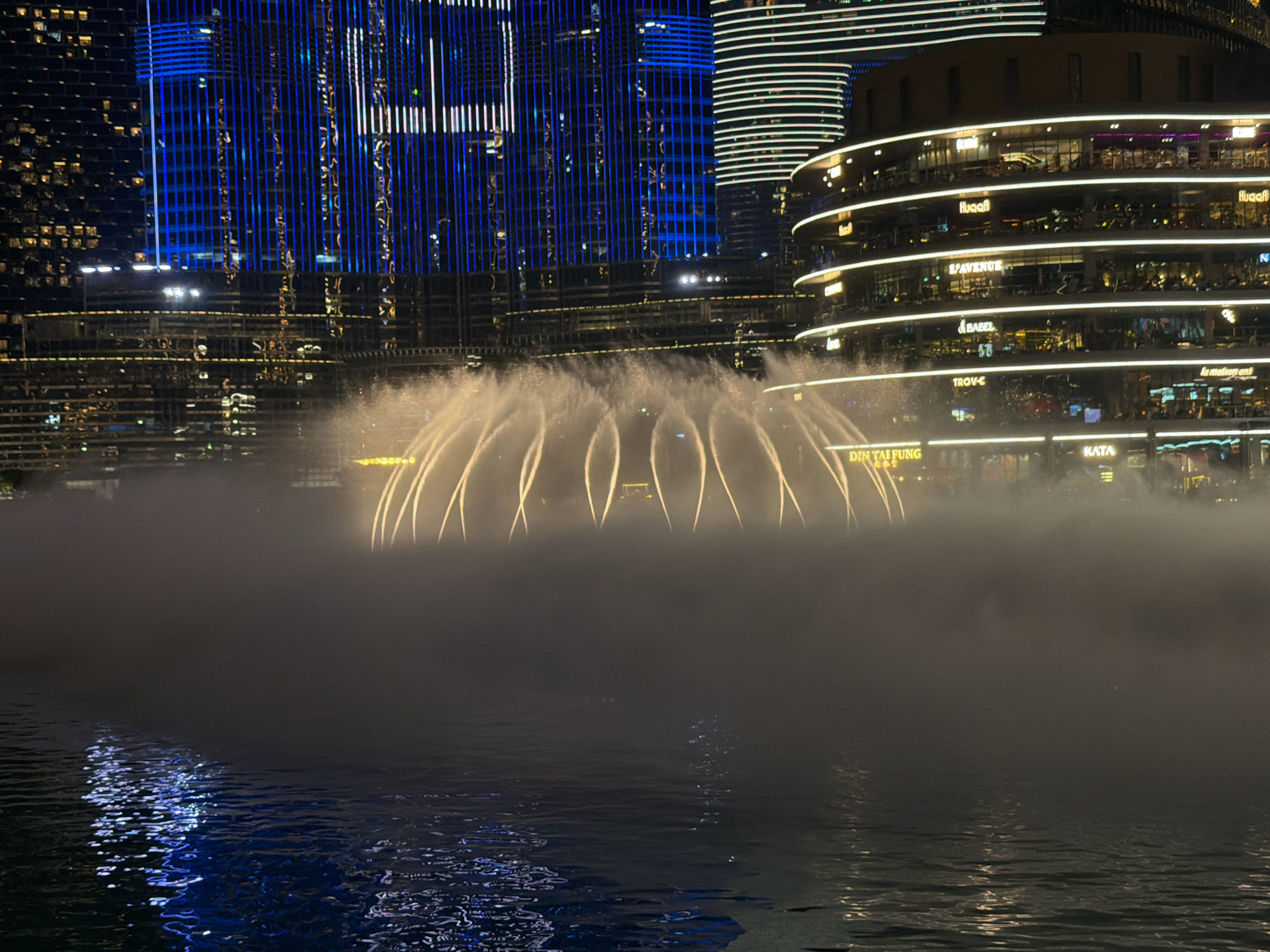 The image shows a nighttime view of the Dubai Fountain, located at the foot of the Burj Khalifa in Downtown Dubai, United Arab Emirates. The fountain is in motion, with illuminated water jets arcing upwards and creating a misty effect. In the background, the distinctive architecture of surrounding buildings is visible, including the Burj Khalifa itself with its blue vertical lighting. The curved facade of a multi-story building, likely a shopping mall or hotel, is also illuminated with white lights and signage, such as "L'AVENUE," "BABEL," "TROY-E," and "DIN TAI FUNG." The water in the foreground reflects the lights from the buildings, creating shimmering patterns. The scene conveys a sense of urban spectacle and entertainment.