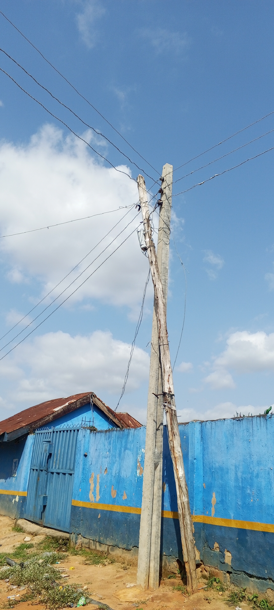 In Jalingo, Nigeria, a scene unfolds under a partly cloudy blue sky featuring a leaning wooden utility pole entwined with a concrete one. The wooden pole appears to be in a state of disrepair, with visible damage and weathering. Attached to these poles are multiple overhead electrical wires, some of which are taut, while others appear slack or damaged. A small electrical transformer is also visible on the wooden pole.
In the background, a low-lying building with a rusted corrugated metal roof is partially visible. This building is bordered by a bright blue, corrugated metal fence, which is marked with a horizontal yellow stripe. The fence shows signs of peeling paint and wear. The ground in the foreground is composed of dirt and scattered green vegetation, along with some visible litter. The overall environment suggests an outdoor setting in a somewhat aged or developing urban area.