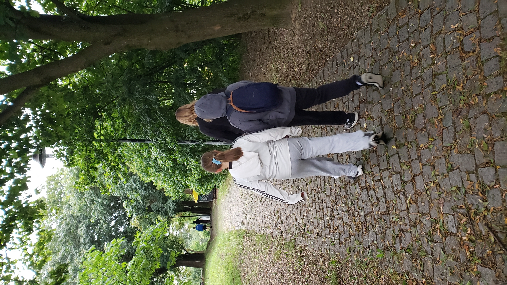 Three people are walking on a cobblestone path through a park. The person in the foreground on the left is a young girl with brown hair in a ponytail wearing a white hooded jacket and grey sweatpants. She is walking with her back to the camera. To her right and slightly behind her is another person, appearing to be a teenager, wearing a grey hooded jacket and dark pants, with a blue backpack. The third person, also appearing to be a teenager, is walking ahead of them, wearing a dark jacket. The path is lined with trees and green foliage. The scene appears to be a public park, possibly within the city of Prešov, Slovakia, given the context of the request.