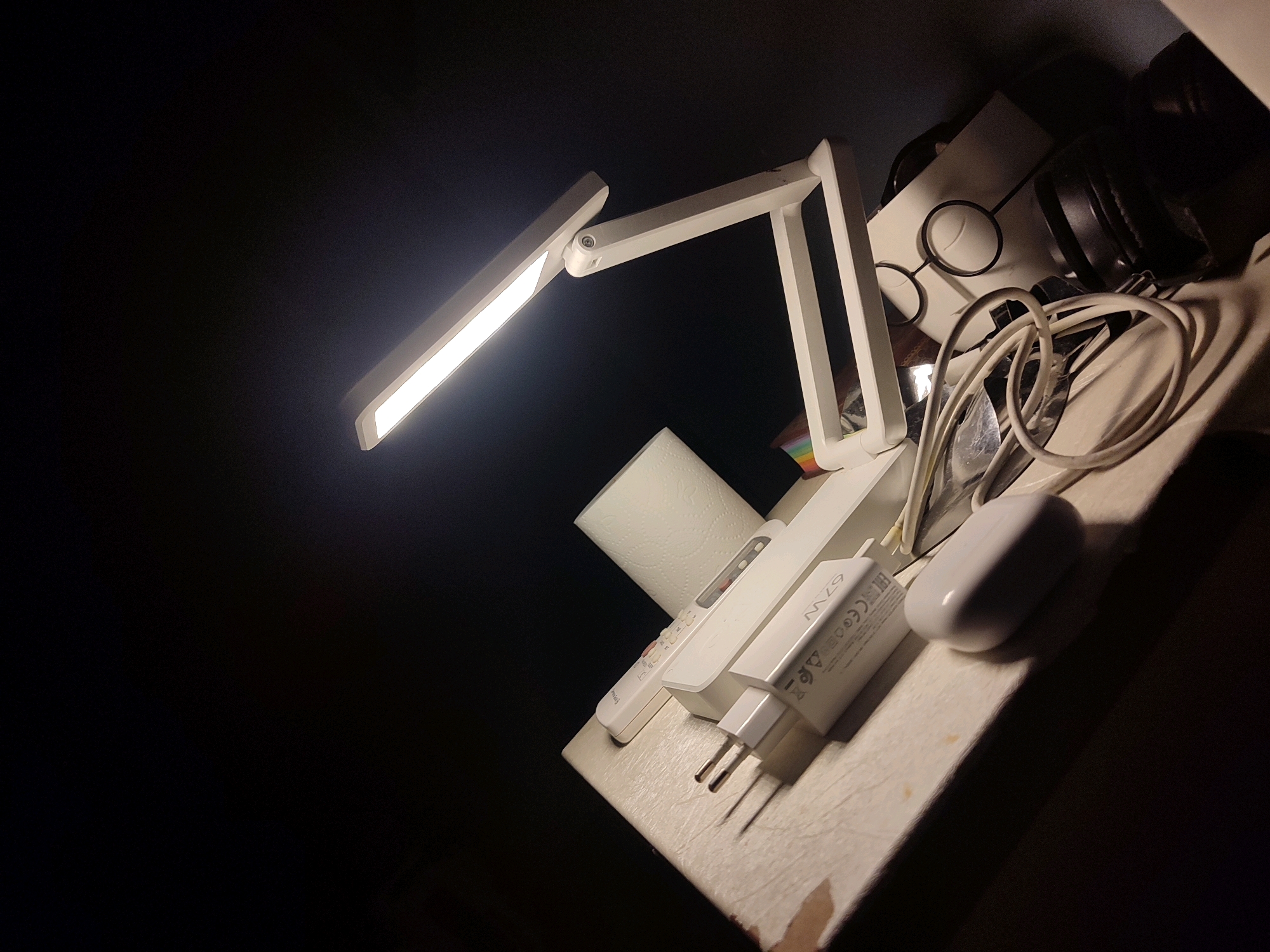 The image captures a dimly lit scene, likely an indoor setting. In the foreground, a white LED desk lamp is illuminated, casting a bright light across a textured surface. To the right of the lamp, a roll of white toilet paper stands upright. Further to the right, several electronic accessories are visible: a white remote control, a white power adapter with "67W" imprinted on it, and a white wireless earbud charging case. Behind these items, a coiled white cable is present. In the background, a pair of black headphones rests on a white surface, alongside a pair of round-framed glasses. The lighting suggests a nighttime or low-light environment. There is no indication of the city of Bechar, Algeria, or any specific geographic location.
