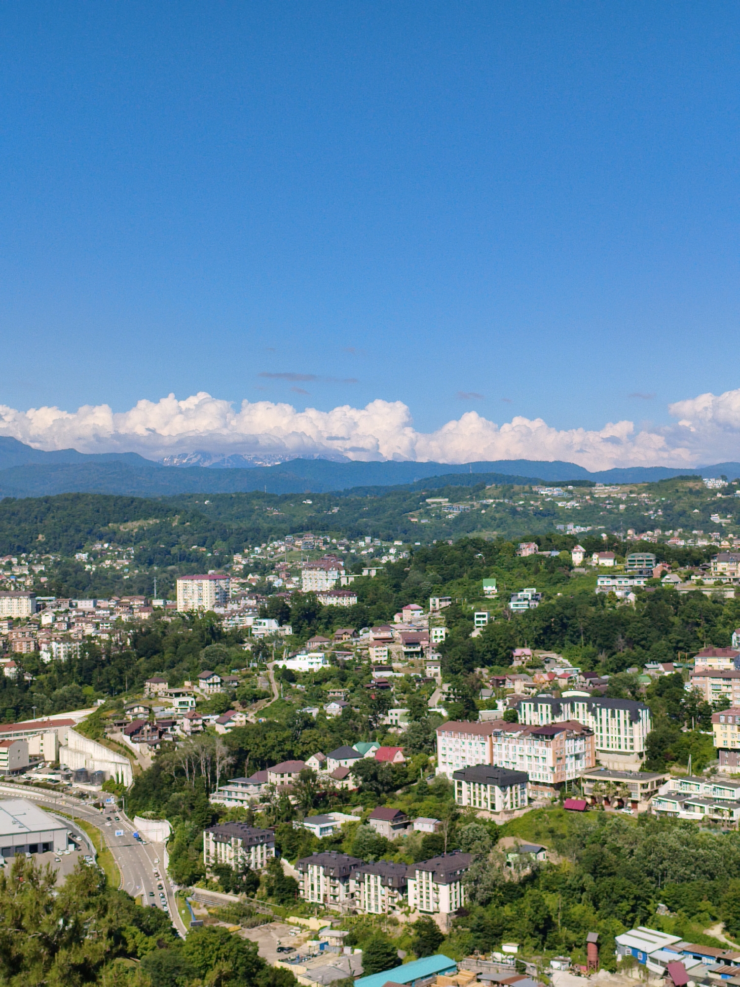 The image depicts a panoramic view of a city nestled on a mountainside, likely in Sochi, Russia, given the context of the provided crops. The cityscape is densely populated with buildings of varying sizes and architectural styles, ranging from multi-story apartment complexes to smaller residential houses, all interspersed with lush green trees and vegetation. A highway curves through the lower part of the frame, with several cars visible on it. In the distance, majestic mountains rise, their peaks partially covered by snow, and the sky above is a vibrant, clear blue, with a few scattered white clouds. The overall scene conveys a sense of urban development harmoniously integrated with a natural, mountainous environment.