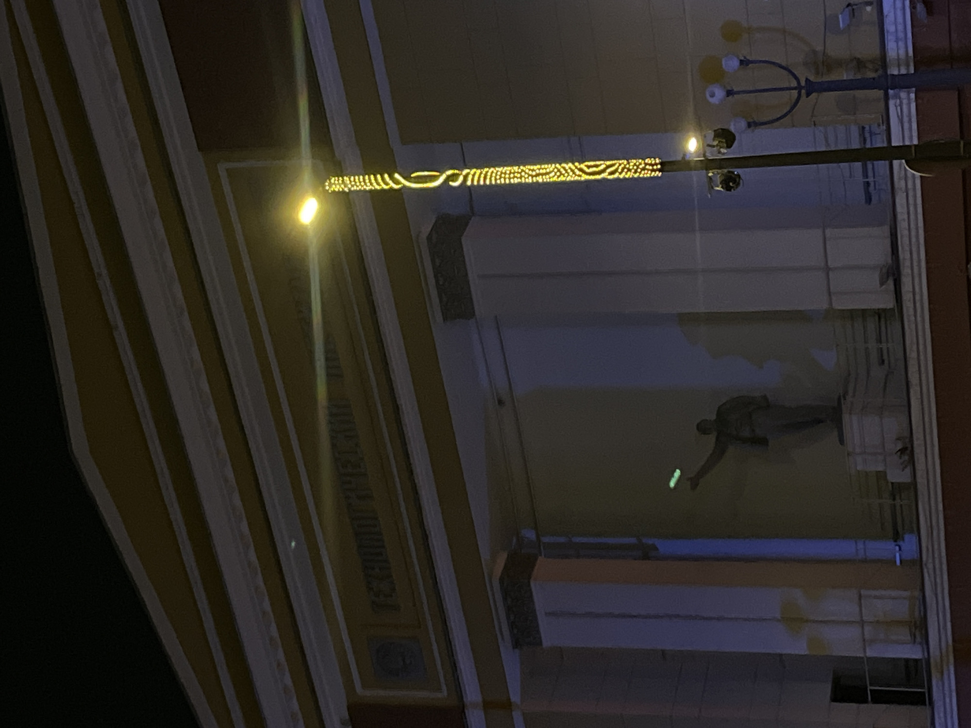 The image captures a nighttime exterior view of a building, rotated 90 degrees clockwise from its natural orientation. The scene is dominated by architectural features and artificial illumination.
The building appears to be a grand structure, possibly with classical or neoclassical elements, featuring a facade composed of vertical light-colored pilasters or columns against a darker, possibly ochre or yellow-brown wall. Above these vertical elements, and spanning horizontally across a portion of the building, are ornate yellow string lights arranged in intricate patterns including swirls, wavy lines, and dotted sections.
On the left side of the image, a very bright, circular yellow light source, likely a streetlamp or spotlight, casts a strong glow. Further to the right, a traditional streetlamp with multiple light globes is visible.
A prominent feature of the building is a recessed niche or alcove where a dark-colored, full-body male statue is situated. The statue stands upright, with its left arm (from the viewer's perspective) extended forward. In the statue's outstretched hand, it holds a distinct, elongated object that emits a bright green glow.
On the building's facade, particularly visible on the darker wall section to the left of the statue, there is embossed or carved text in Cyrillic script. The visible portion clearly reads "ТЕАТР ИМ. ГЕЛ..." (TEATR IM. GEL...), indicating it is a theatre named after an individual or entity starting with "Gel".
The overall impression is that of a public or institutional building, likely a theatre, adorned with decorative lighting at night, and featuring a notable statue with an unusual glowing prop.