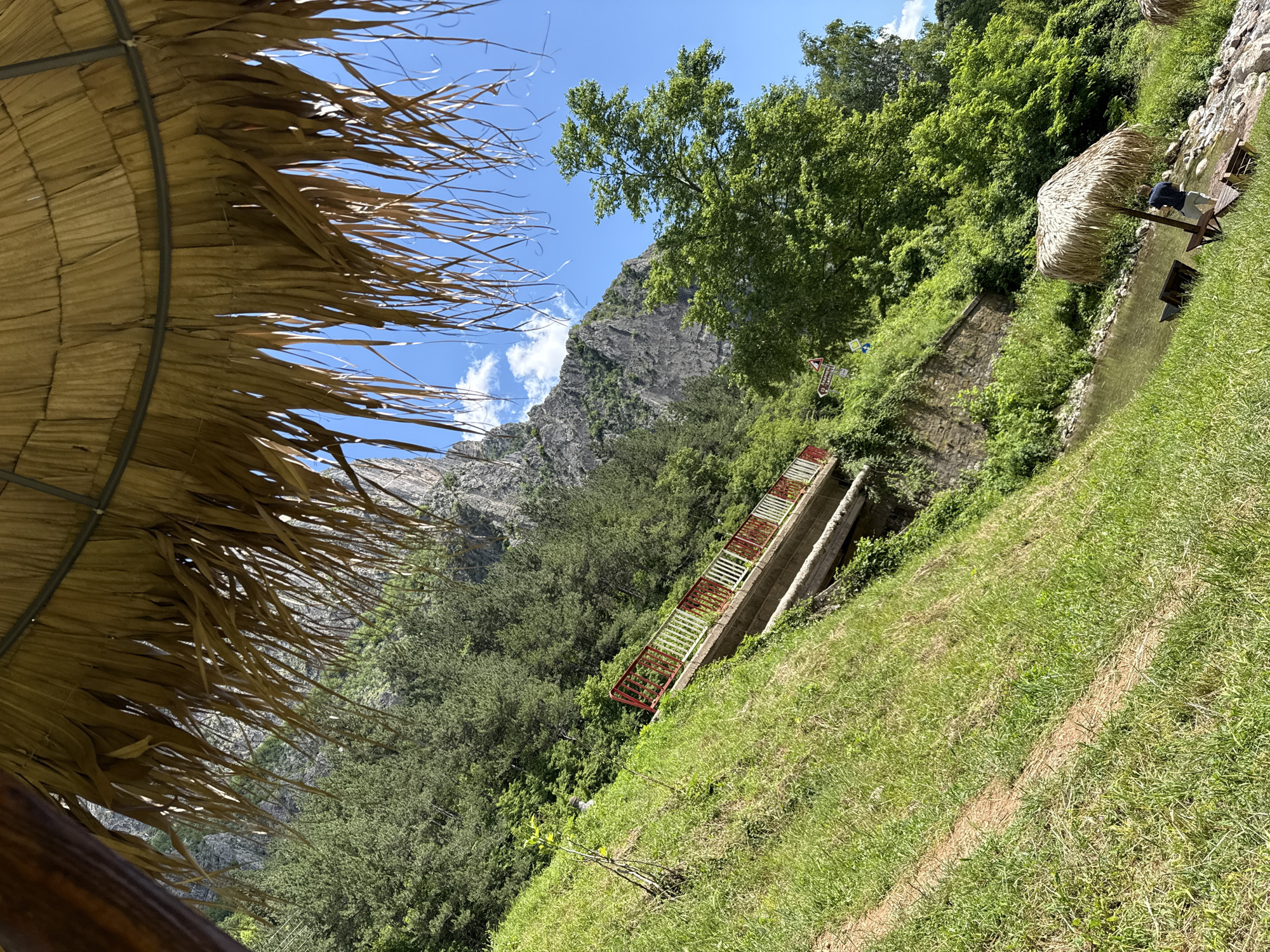 The image captures a scenic view of a natural environment, possibly a park or a riverbank, with a mountainous backdrop. In the foreground, a thatched umbrella provides shade, with two empty lounge chairs positioned on a grassy area next to a body of water. Further back, a small bridge with red and white railings spans across a slope covered in lush green grass and vegetation. The bridge is situated on the side of a steep incline, leading up towards a rocky mountain face. Dense green trees and foliage cover the slopes and mountainside, with a clear blue sky dotted with white clouds overhead. The overall impression is one of a tranquil outdoor setting, likely a popular spot for recreation and relaxation. The location is identified as Zgorigrad, Bulgaria, suggesting it's a natural landmark or recreational area within or near the city.