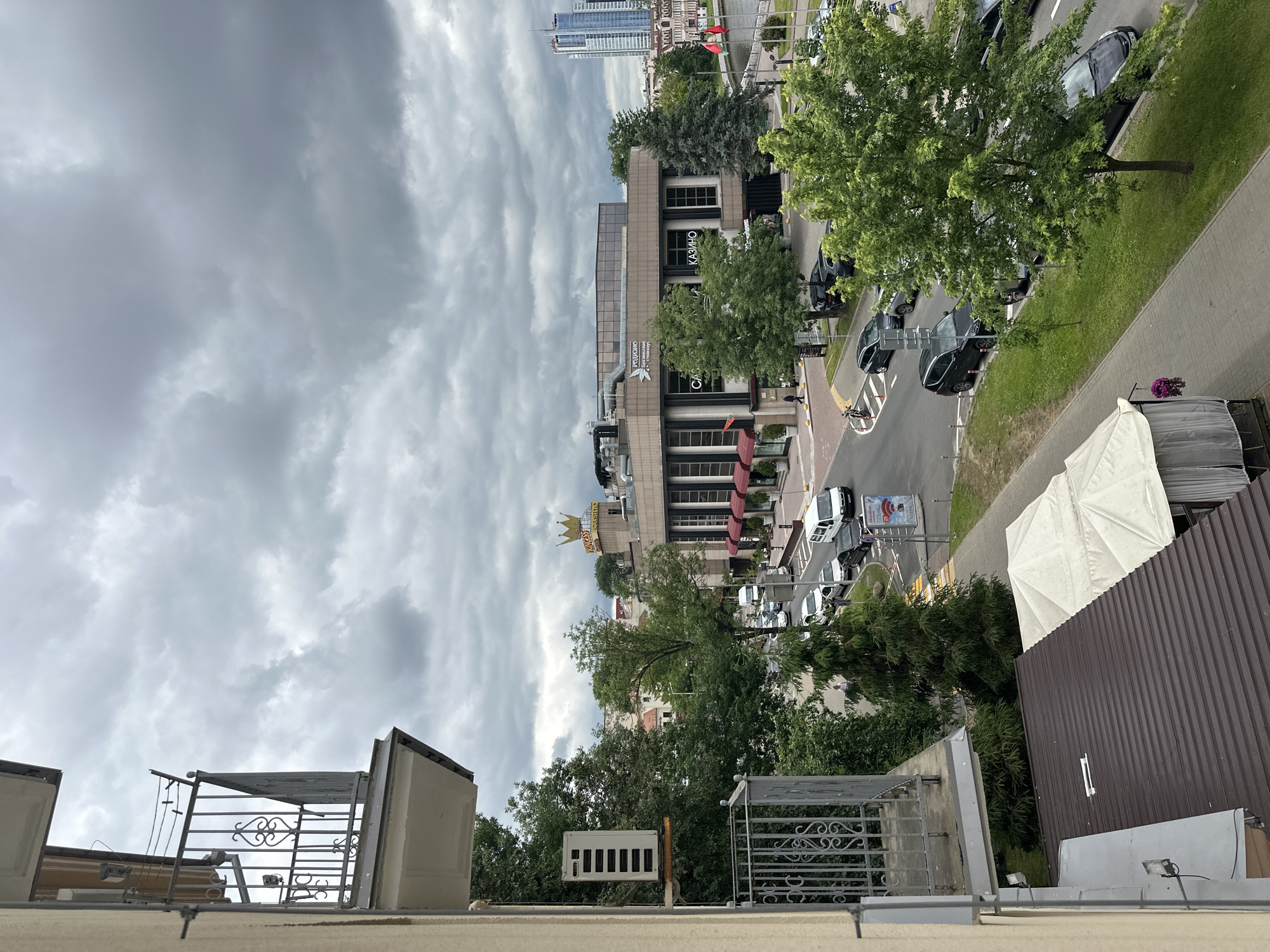The image depicts a street view in Minsk, Belarus, likely taken from an elevated vantage point such as a balcony. The sky is heavily overcast with dark, textured clouds. In the foreground, the side of a building is visible, with a balcony featuring a metal railing and an air conditioning unit mounted on the wall. Below the balcony, a dark metal roof with a white awning or tent is partially visible.
The main focus of the image is a street scene with several cars, including white vans, black sedans, and other vehicles, moving or parked along the road. A large, circular building with a prominent golden crown logo on top is situated in the mid-ground. The building appears to be a casino or entertainment venue, with signage that includes "KING'S CASINO" and Cyrillic text. Adjacent to this building, another structure with large windows and red awnings is visible. Trees with lush green foliage line the street and surround the buildings, indicating a temperate season. In the distance, modern skyscrapers can be seen against the cloudy sky, suggesting an urban environment. A body of water, possibly a river or canal, is also present in the background, with a bridge or embankment alongside it. A pedestrian can be seen walking on the sidewalk.