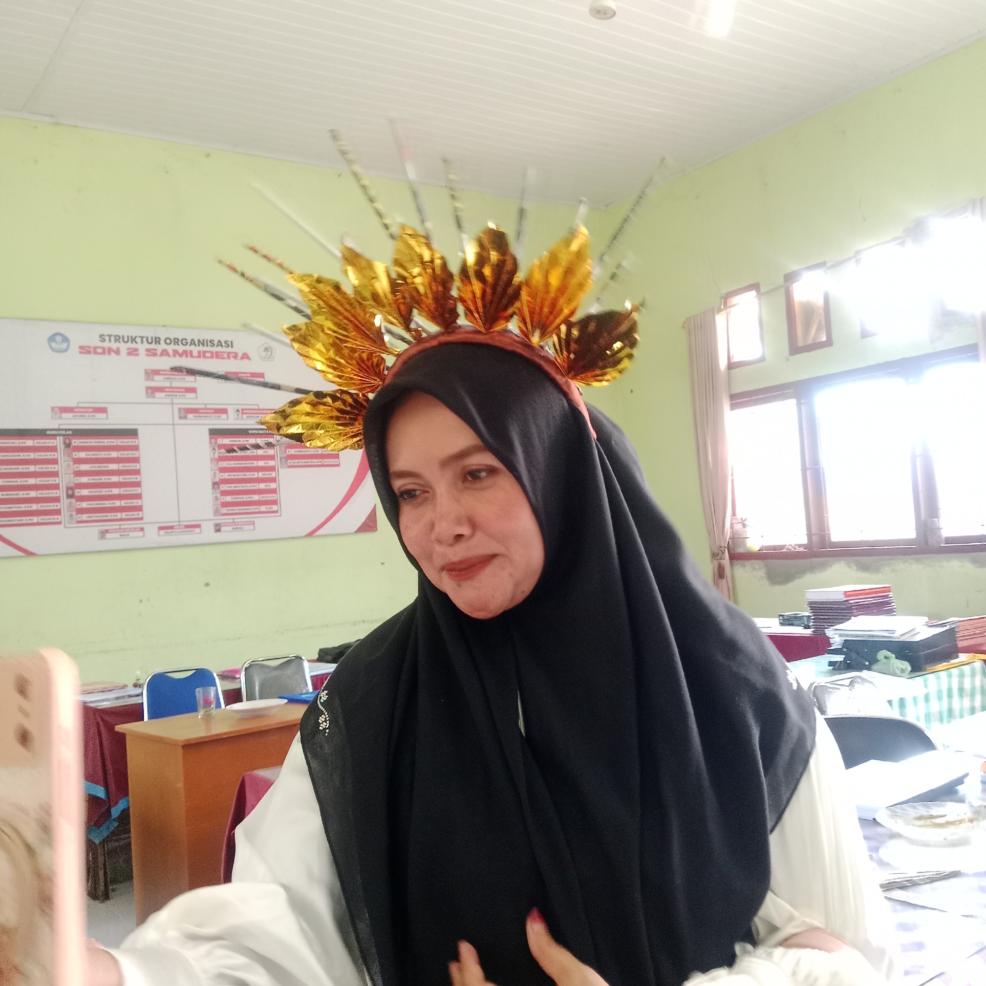 The image shows a woman wearing a black hijab and a golden headdress, possibly a costume, in what appears to be a classroom or office setting. She is holding a light pink smartphone, suggesting she is taking a selfie. In the background, a whiteboard with a "STRUKTUR ORGANISASI SON 2 SAMUDERA" chart is visible, indicating the location is likely "SON 2 SAMUDERA," a school. There are windows with curtains, desks, chairs, and stacks of papers on tables, suggesting an educational environment. The lighting is bright, with sunlight streaming in from the windows, creating some glare. The woman's face is visible, looking towards the camera. The room's walls are painted a light green, and the ceiling is white with visible paneling.