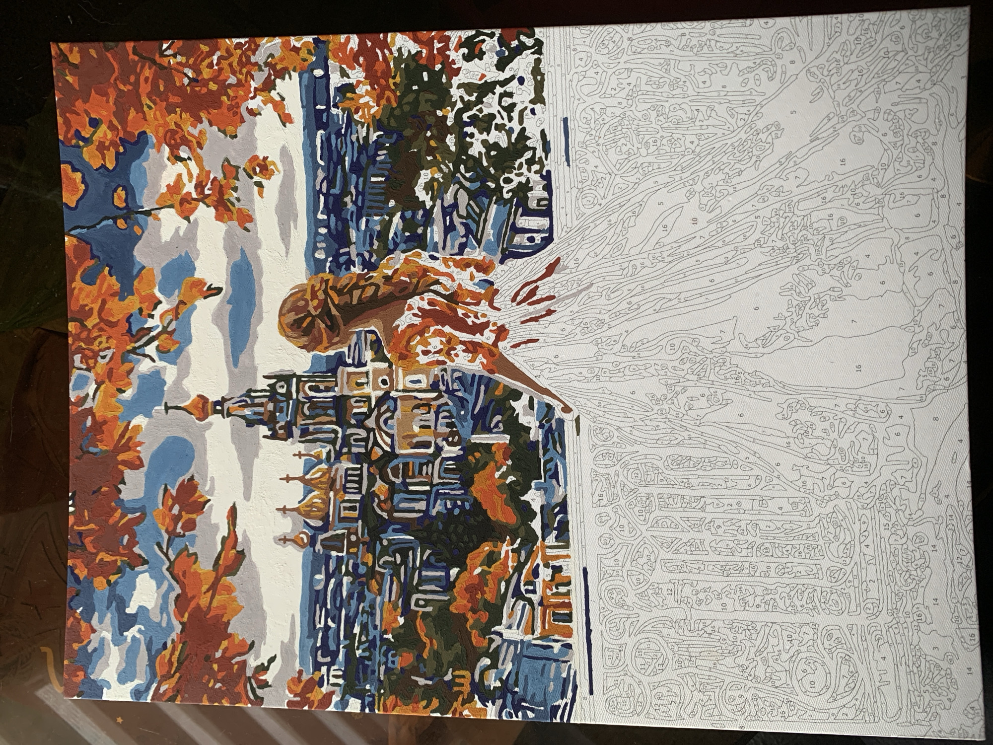The image displays a partially completed paint-by-number canvas depicting a panoramic view of the city of Chaikovsky, Russia, during autumn. A lone female figure, seen from the back, stands in the foreground, looking out over the cityscape from an elevated vantage point, possibly a balcony or terrace. Her hair is styled in a braid, and her upper body, including the shoulders and bodice of a long dress, is painted in shades of orange and brown. The lower portion of her flowing white dress, extending to the bottom of the canvas, is unpainted and marked with numerous numbered sections for the paint-by-number activity.
The background features the city of Chaikovsky under a partly cloudy sky with patches of blue and grey. Prominent in the mid-ground are several buildings exhibiting classic Russian architectural characteristics, including golden onion domes and distinct spires, likely representing churches or significant historical structures. The surrounding urban landscape is depicted with a mosaic of blue, grey, green, and brown tones, indicating various buildings and some green spaces. Vibrant autumnal foliage, characterized by bold blocks of orange and red leaves, frames the top and sides of the scene, adding depth and seasonal context. The foreground's railing or balustrade, like the woman's dress, remains unpainted and shows a network of numbered segments outlining its intricate design.