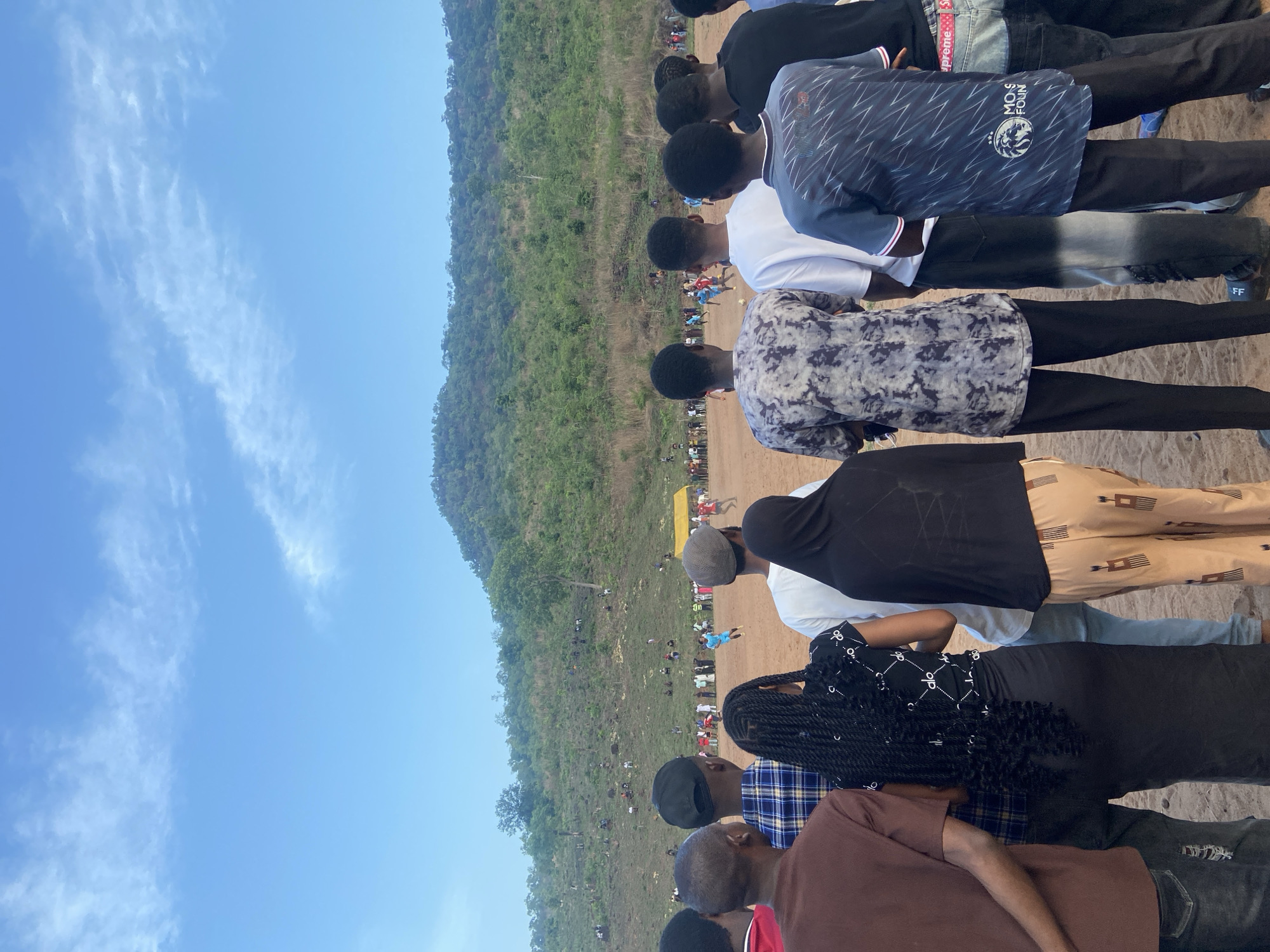 A ground-level, outdoor shot shows a line of people, mostly young men, overlooking a field in what appears to be a mountainous area in Lokoja, Nigeria. The focus is on the backs of these people, who are dressed in casual clothing, including T-shirts, button-down shirts, and pants. The crowd appears to be observing an event taking place on the field, which has a dirt surface and is lined with spectators. Behind the field is a hill covered in green vegetation. The sky is blue with light clouds.