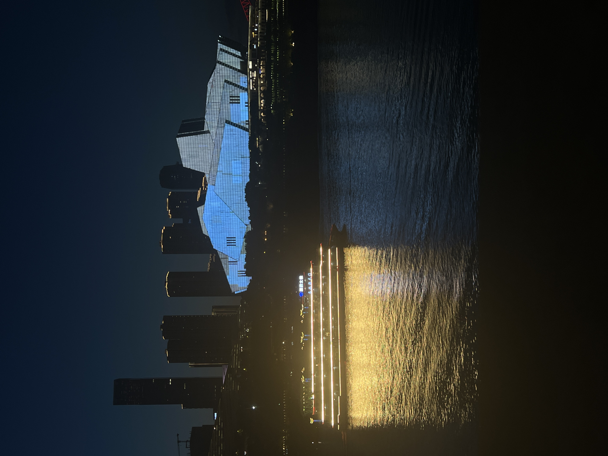 A nighttime panoramic view captures the cityscape of Yuzhong District, China, with the Yangtze River in the foreground. A multi-decked passenger boat, illuminated by numerous golden lights, glides across the dark water, its reflection shimmering on the surface. In the background, a modern architectural marvel, likely a performance venue, is brightly lit with geometric patterns of blue light. Behind this, several silhouetted skyscrapers pierce the night sky, their upper floors occasionally catching faint light. The overall scene is one of urban vibrancy and nocturnal beauty.