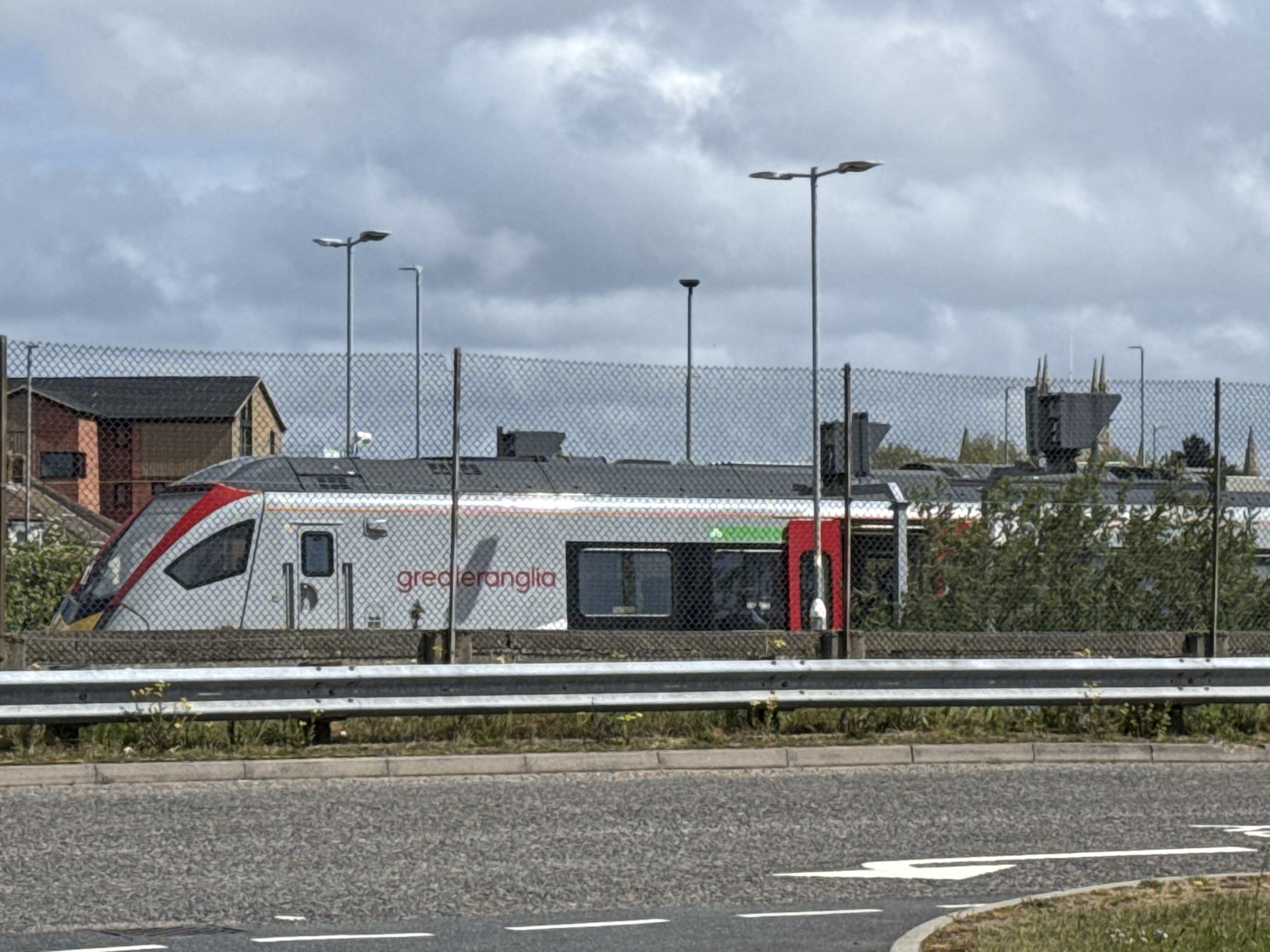The image shows a Greater Anglia train behind a chain-link fence in Great Yarmouth, United Kingdom. The train is silver with red and green accents, visible through the fence. In the background, there's a residential building and streetlights. A grey road with white markings is in the foreground. The sky is overcast. The fence obscures some of the train and background details. A metal crash barrier stands between the road and the fence.