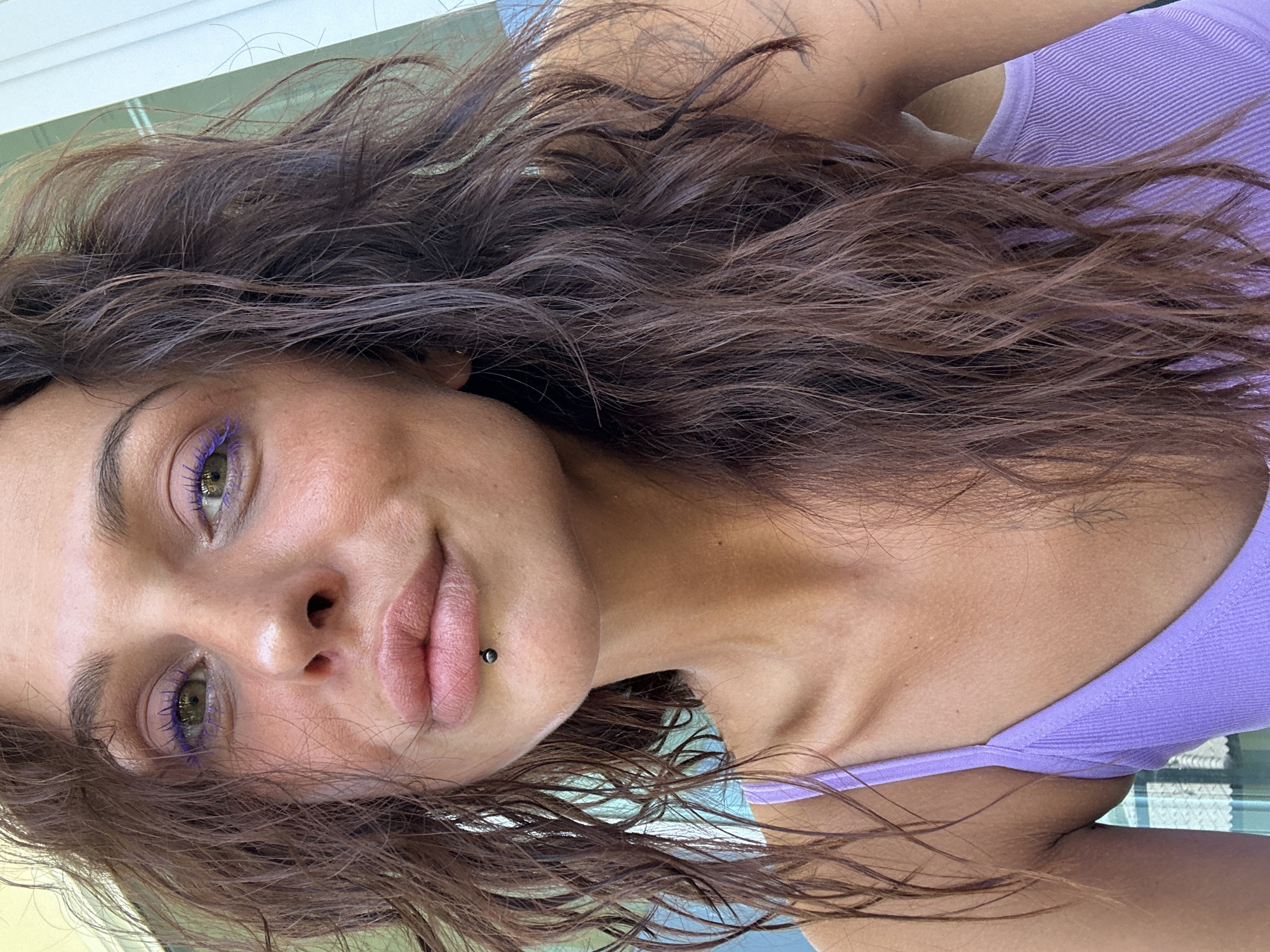 A close-up, head-and-shoulders portrait of a woman is captured outdoors, likely in Ko Samui District, Thailand, judging by the tropical lighting and possibly the faint background elements. She has dark, wavy hair that frames her face, and her eyes are accentuated with vibrant purple eyeliner. A small silver stud is visible in her lower lip. She is wearing a lavender-colored tank top. The lighting suggests it is daytime, and the overall impression is casual and relaxed.