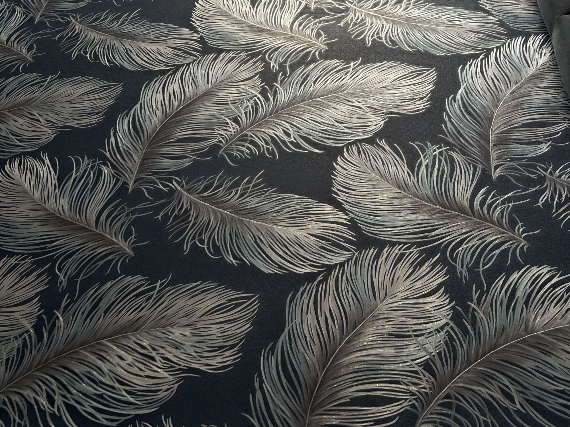The image shows a dark wallpaper with a repeating pattern of stylized feathers. The feathers are depicted in shades of grey, silver, and muted teal, with a subtle shimmer. They are arranged in a scattered, organic manner across the dark background. The image is a close-up, likely of a wall in a room. There is no visible indication of the city of Ust'-Katav, Russia.