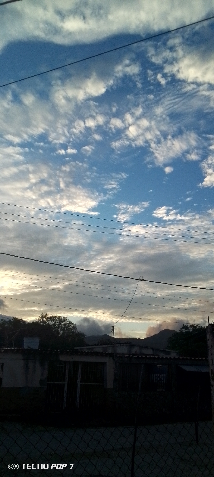 The image captures a view of the sky and a row of buildings in Altagracia, Venezuela. The sky is a prominent feature, filled with a mix of blue sky and scattered white clouds, some of which are wispy and others more solid, with a hint of sunlight breaking through in the distance. Several dark electrical wires are strung horizontally across the frame.
In the foreground, a low-rise building with a tiled roof is visible. It appears to be a residential or commercial structure with barred windows and doorways. The lower portion of the image shows a dark chain-link fence. Beyond the building, a line of trees and hills can be seen against the horizon, shrouded in a light haze. The overall lighting suggests it is either early morning or late afternoon, given the soft light and the cloud cover. There are no people clearly visible in the image. The "TECNO POP 7" watermark is present in the bottom left corner, indicating the device used for capture.