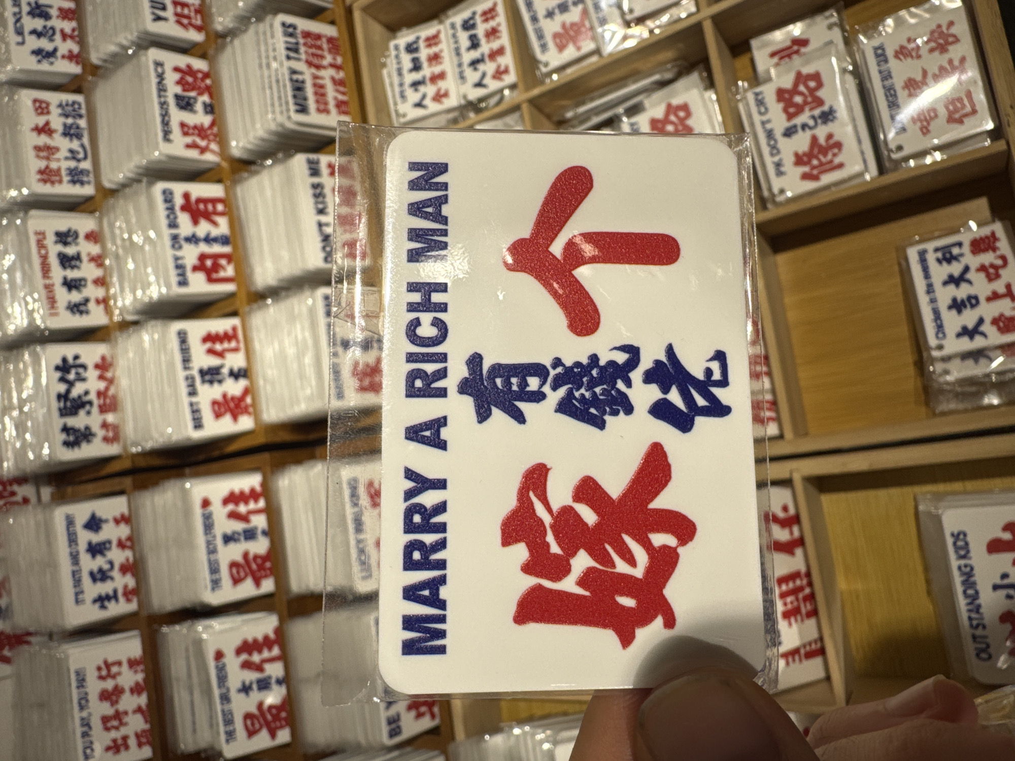 A person is holding a packaged card in a shop or market, likely in the Liwan District, China, given the presence of Chinese characters on many cards. The card the person holds is white with blue text reading "MARRY A RICH MAN" vertically. Below this text, there are larger red Chinese characters. Other cards are visible in a wooden compartmentalized display case, some with English phrases like "BABY ON BOARD", "DON'T KISS ME", "MONEY TALKS", and "PERSISTENCE" along with Chinese characters.