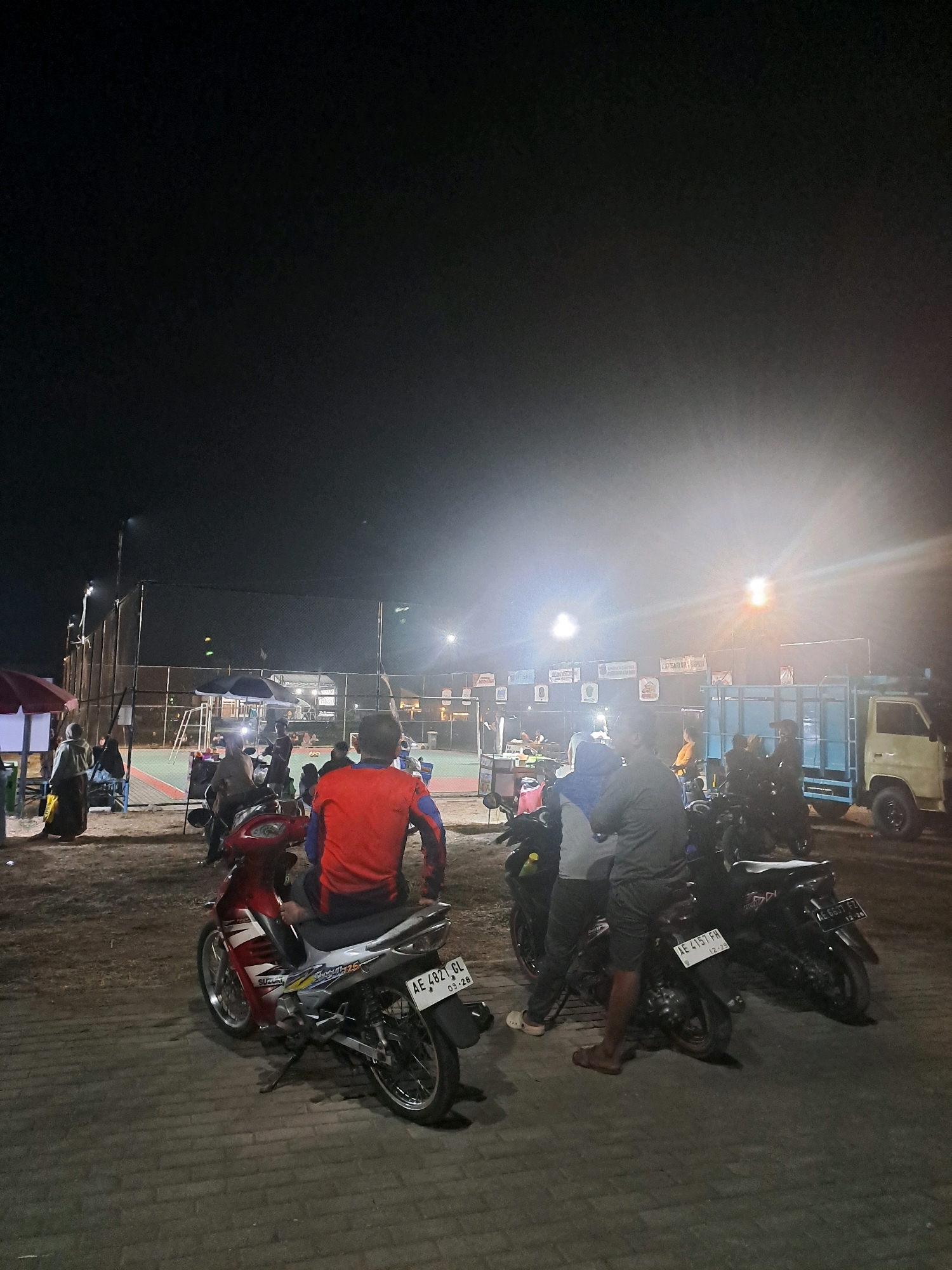 The image depicts an outdoor nighttime scene at what appears to be a sports facility, possibly a badminton or tennis court, located in Sumberejo, Indonesia. The lighting is primarily from bright, overhead floodlights, casting a significant glare. In the foreground, several motorcycles are parked on a paved walkway. A person in a red jacket is seated on a red and silver motorcycle, facing away from the camera. To the right of this motorcycle, two individuals are standing next to a black motorcycle, one wearing a grey top and shorts. Further in the background, a light blue truck with a light yellow cab is parked. Beyond the court, there are various tents and structures, some with signage. Several other people are visible in the background, some near the court and others gathered around stalls or seating areas. The overall atmosphere suggests a public gathering or event taking place after dark.