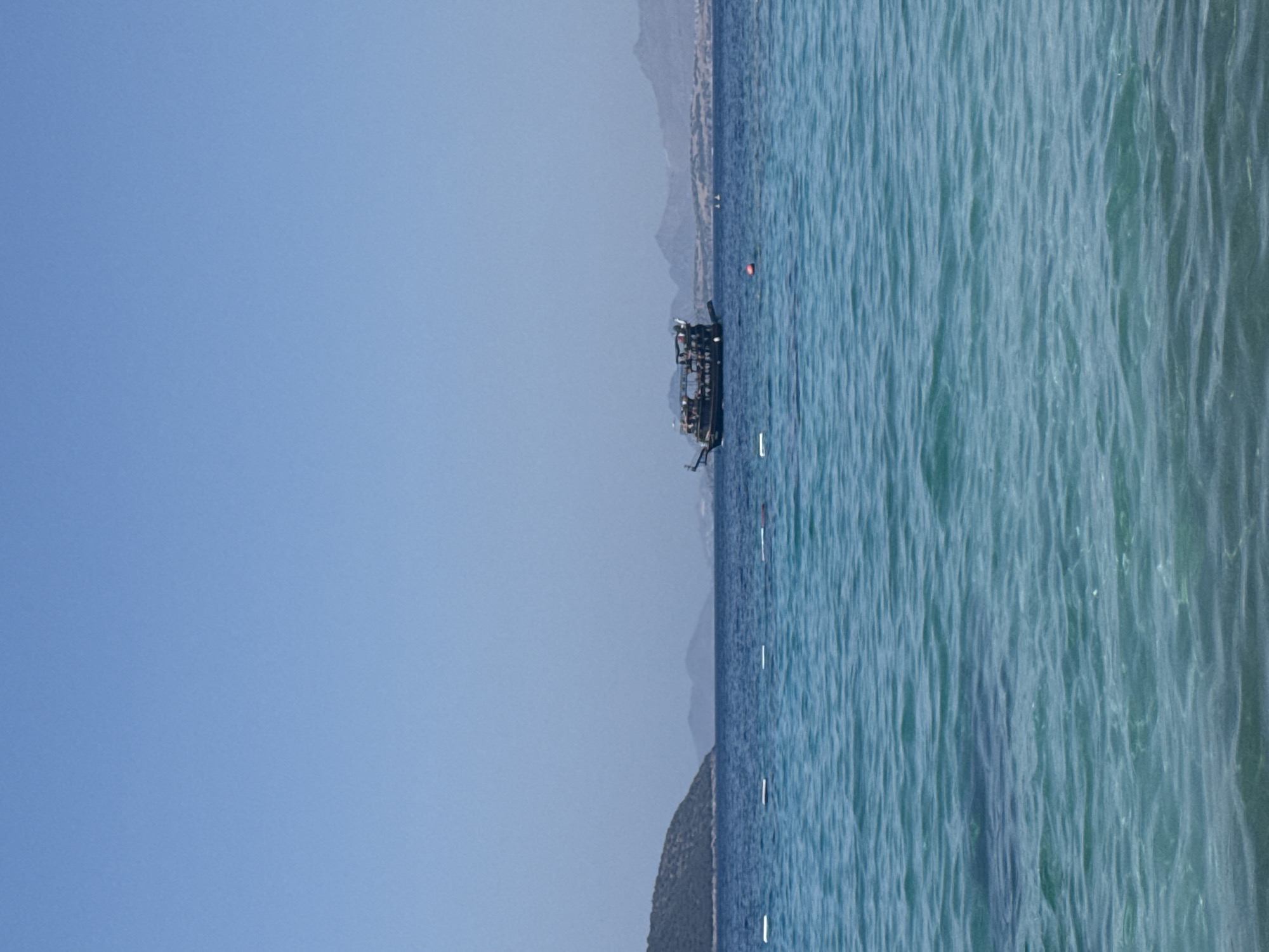 A side view of a large, dark-colored boat with multiple decks is sailing on a calm, blue sea. The boat has a distinct shape, appearing to be a traditional Turkish gulet or a similar type of wooden vessel. Several people are visible on the decks of the boat, some standing and others seated. The boat is positioned in the middle ground of the image, with a clear blue sky above and a lighter blue ocean in the foreground. In the background, a green, tree-covered island is visible to the left, and a range of hazy mountains stretches across the horizon. The overall scene depicts a sunny day at sea, likely near the coast of Bodrum, Turkey, given the presence of traditional boats and the characteristic landscape.