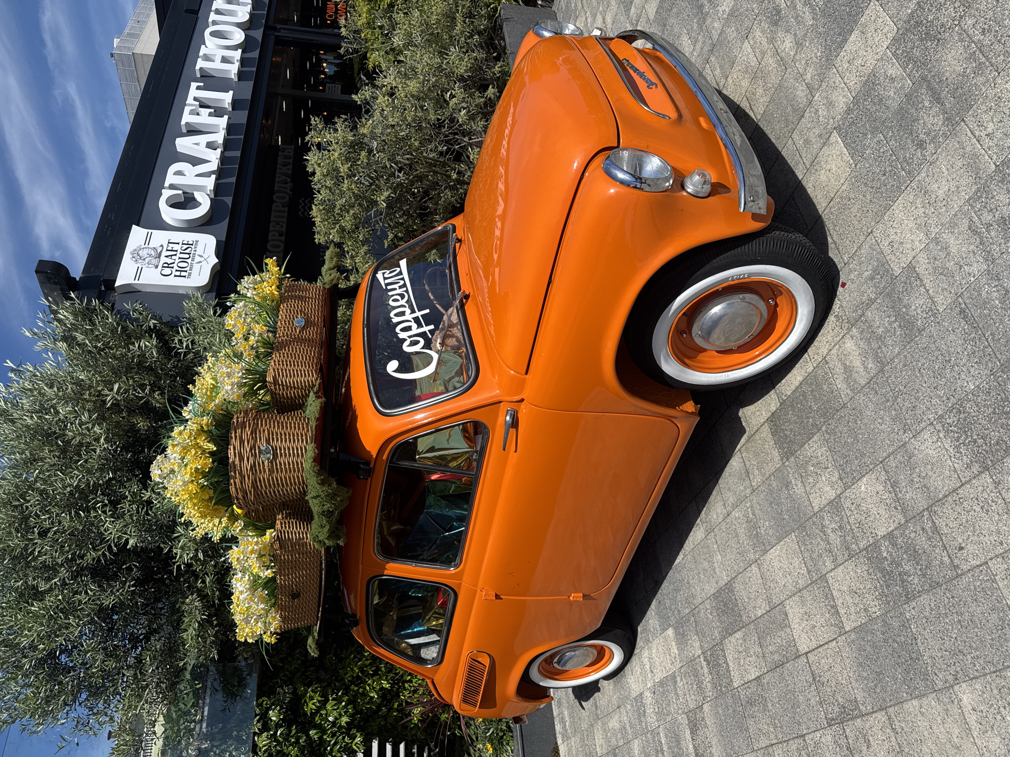 An orange vintage car, likely a Zaporozhets given the Cyrillic text "Запорожець" visible on its front, is parked on a paved surface under a bright, sunny sky in Sochi, Russia. The car is adorned with multiple wicker baskets filled with an abundance of yellow and white flowers, possibly daffodils or similar spring blooms, arranged on its roof rack. The vehicle features classic design elements, including white-wall tires and chrome accents, and has the word "Сорренто" (Sorrento) visible as a decal on its windshield.
In the background, a dark-colored building with large white lettering proudly displays the name "CRAFT HOUSE." Beneath the main sign, a crest-like logo further clarifies the establishment as "CRAFT HOUSE THE BEST BEER & WINE." Other Cyrillic text is visible on the building facade, including "ОРЕПРОДУКТЫ" (likely a misspelling or partial word for "nut products" or similar retail goods) and "СУШИ" (Sushi), indicating a diverse offering. Green foliage, including bushes and what appears to be an olive tree, frames the scene, adding natural elements to the urban setting. The ground is paved with rectangular grey and lighter-toned tiles. The clear blue sky with sparse clouds suggests a pleasant daytime setting. The car and its floral display appear to serve as a decorative or promotional feature for the "CRAFT HOUSE."