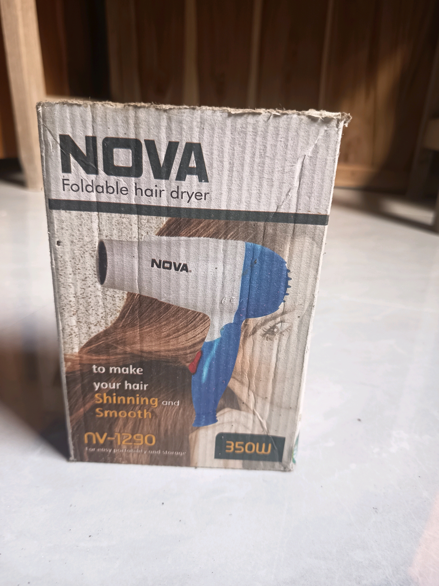 A cardboard box for a "NOVA Foldable hair dryer" sits on a tiled floor. The box has an image of a white and blue hair dryer and text indicating "to make your hair Shinning and Smooth," model number "NV-1290," and "350W." The box appears worn, with visible creasing and slight damage to its edges. The background consists of wooden planks, possibly part of a wall or furniture. The lighting suggests an indoor environment. There are no people or any specific indicators of the city of Sidorejo, Indonesia.