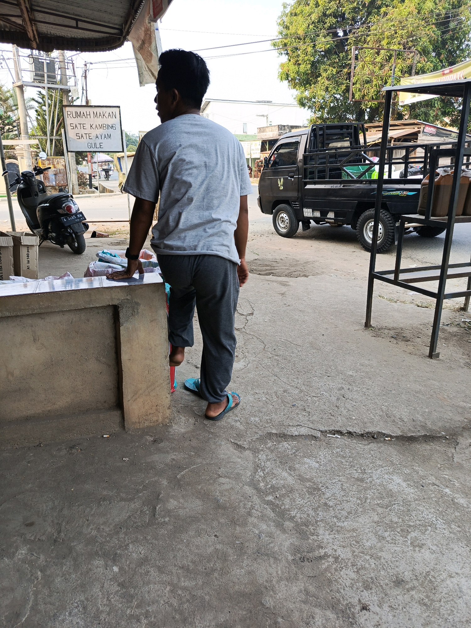 The image depicts an outdoor scene in front of a "RUMAH MAKAN SATE KAMBING SATE AYAM GULE" (Goat Satay, Chicken Satay, Gule Restaurant) in Megang Sakti I, Indonesia. A man with dark hair, wearing a gray t-shirt and gray pants, stands with his back to the camera, facing a concrete counter. He appears to be resting his left hand on the counter and has his right leg extended behind him, resting on a blue and black flip-flop. His left leg is bent and is also in a blue and black flip-flop.
Behind the man, to the left, is a dark-colored scooter. To the right of the scooter, the restaurant's sign is visible, listing the menu items. Further to the right, a black pick-up truck is parked. The truck has a metal cage attached to its rear. In the foreground, the ground is rough concrete with some debris. In the background, there are trees and buildings, suggesting an urban or semi-urban environment. The overall lighting suggests it is daytime, possibly late afternoon due to the warm tones.