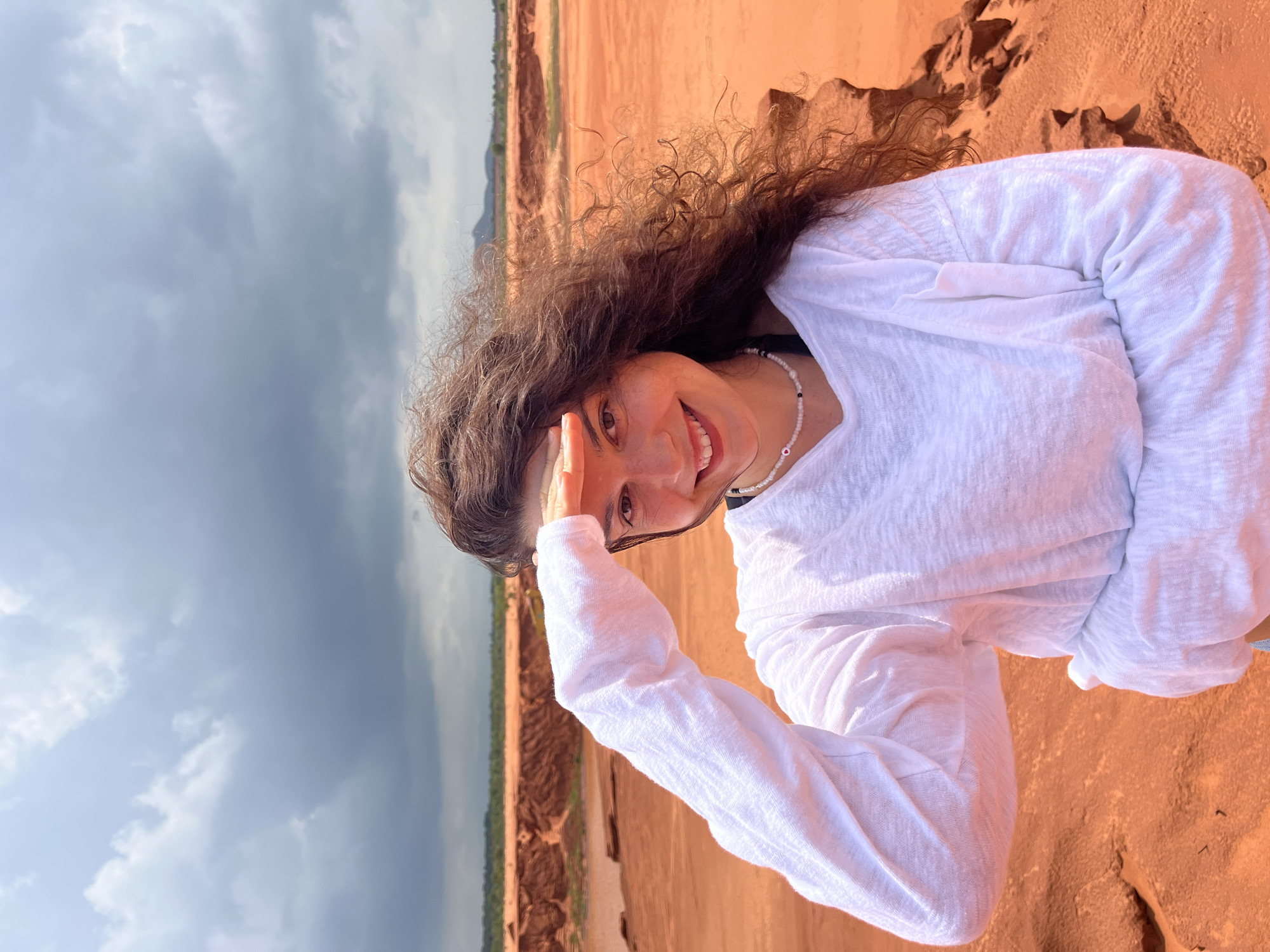 In Mui Ne, Vietnam, a woman with long, curly brown hair stands in a sandy, arid landscape under a cloudy sky. She is wearing a white long-sleeved shirt and a beaded necklace. Her right hand is raised to shield her eyes from the sun as she looks towards the viewer with a smile. The background features red sand dunes and sparse vegetation, suggesting a desert-like environment.