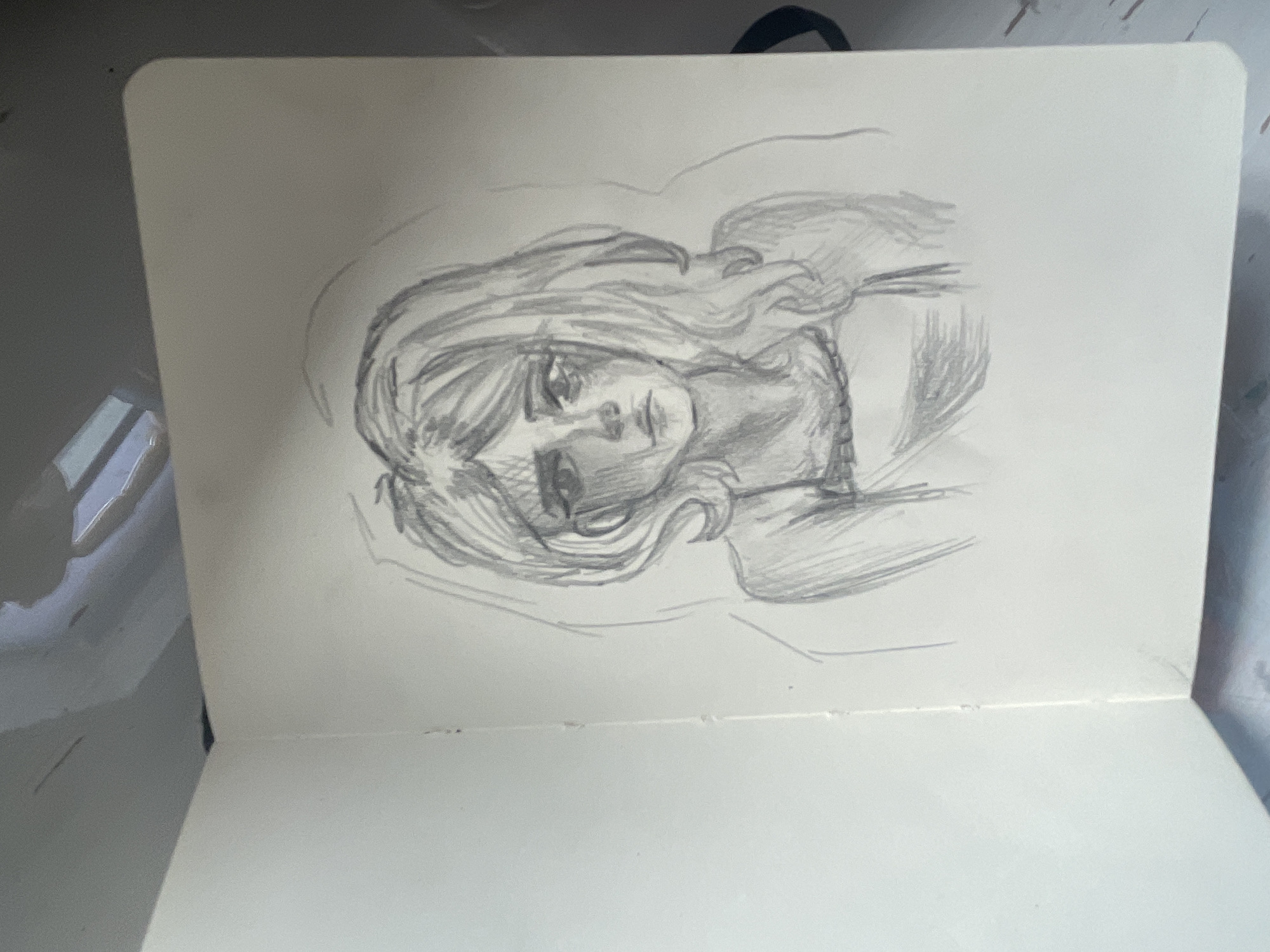 A pencil sketch of a young woman with wavy hair is depicted in a sketchbook. She has a somber expression and is wearing a simple dress. The background is lightly sketched, giving a sense of depth. The sketchbook is open, and the page on the right contains the drawing. The environment appears to be indoors, with a hint of reflections on a surface behind the sketchbook, suggesting it might be placed on a table or desk. The location of Мухавецкі сельскі Савет, Belarus is not discernible from the image.