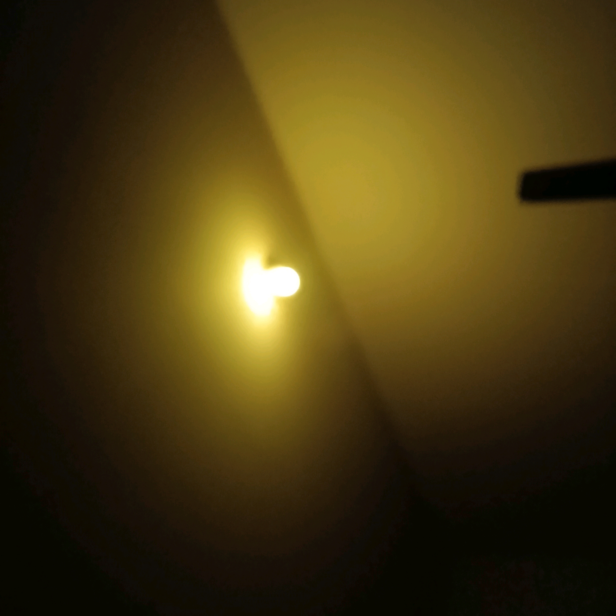 The image depicts a dimly lit room with a strong yellow light source emanating from the left side, creating a bright, blurred circular glow. A dark, elongated object, possibly a shadow or a physical object, is visible in the upper right quadrant of the image. The background is dark and indistinct, with gradients of yellow and brown suggesting a wall or ceiling. The lighting conditions indicate a low-light environment, possibly artificial. There are no people or discernible activities taking place. The image does not contain any visual cues to determine its specific location within Bengaluru, India, or any other geographical context.