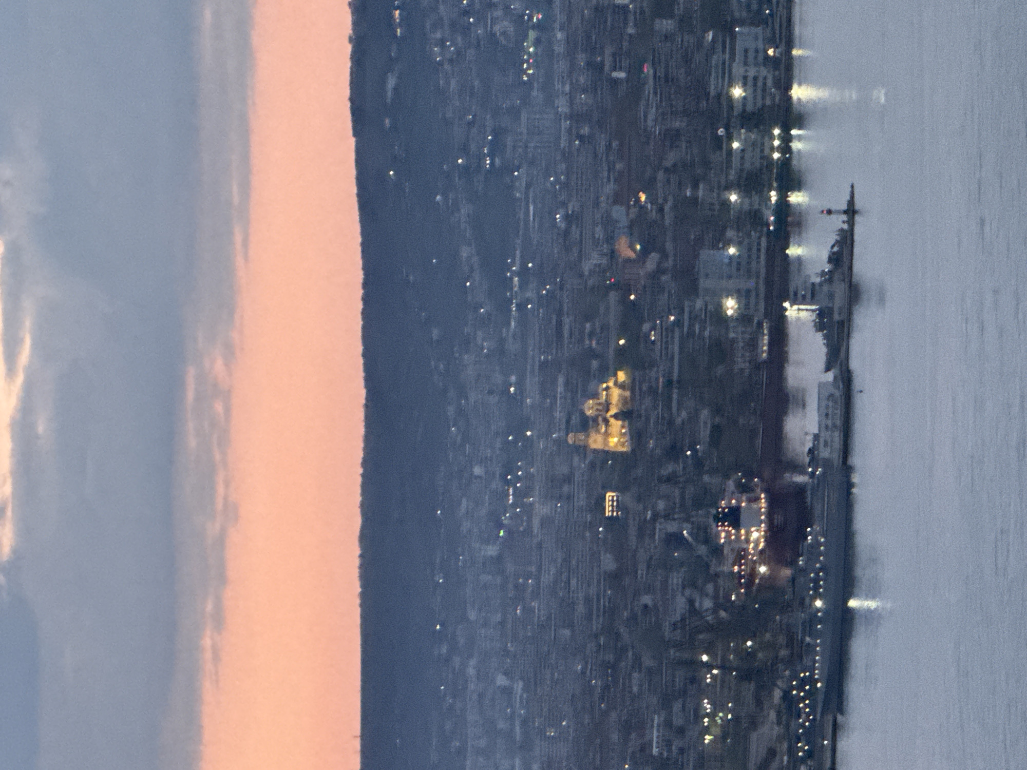 This image presents a panoramic view of a city at dusk or early night, looking across a calm body of water. The scene is illuminated by both the fading natural light and the numerous artificial lights of the urban area.
The sky, occupying the upper portion of the frame, displays a beautiful gradient of colors, transitioning from a light blue-grey at the top to a warm peach and orange closer to the horizon, indicative of either sunset or sunrise. Below the sky, a dark, undulating silhouette of a mountain or high ground forms a distinct horizon line, behind which the city is situated.
The city itself is a vibrant tapestry of lights, stretching across the landscape and appearing to ascend the slopes of the distant landmass. A prominent, brightly illuminated building with golden domes stands out in the mid-ground of the city, strongly identifiable as the Dormition of the Theotokos Cathedral, a well-known landmark in Varna, Bulgaria. Numerous other lights dot the urban expanse, highlighting buildings, streets, and various structures.
In the foreground, a large body of water, likely a bay or the sea, reflects some of the city's glow. The water appears tranquil, with subtle ripples. A sizable vessel, possibly a naval ship or a cargo ship, is clearly visible docked or anchored in the water near the city lights, contributing its own illumination to the scene.
No specific activity or event is taking place, and no visible text is discernible. The overall atmosphere conveyed is one of serene beauty, capturing the transition from day to night in a bustling port city. The distinct architecture of the cathedral and the presence of a large port strongly suggest the location is Varna, Bulgaria.