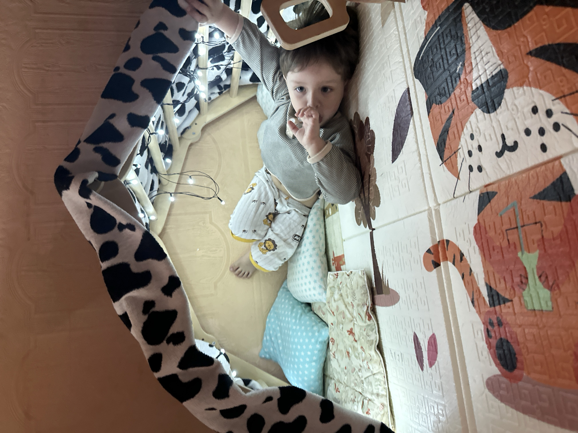 A young child is lying on their back inside a blanket fort, which is decorated with string lights. The fort is constructed using a blanket with a cow-print pattern. The child is wearing pajamas with a lion print. There are several pillows and a small quilted mat inside the fort, which is set up on a floor mat with a tiger design. The background appears to be a wall with wooden paneling. The overall scene suggests a cozy, indoor play area, possibly a child's bedroom. The image does not contain any information that would allow for an identification of its location in Vladivostok, Russia.