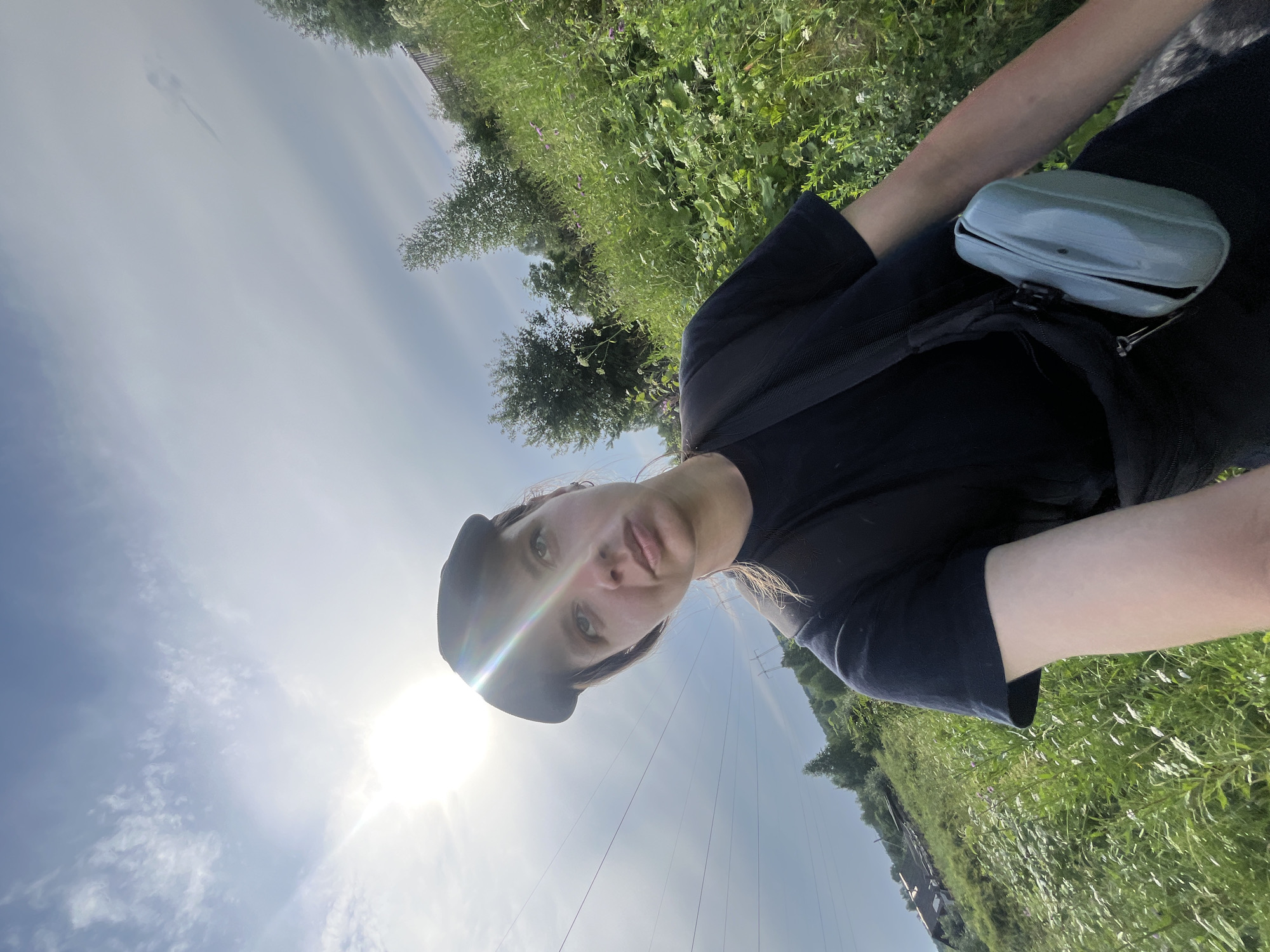 A young woman wearing a black t-shirt, black cap, and a light blue fanny pack is taking a selfie outdoors. She has light skin and light hair pulled back. The sun is bright and high in the sky, creating lens flare. The background is a grassy hillside with trees and some distant houses. Power lines are visible in the background. The location is the city of Nizhny Tagil, Russia.