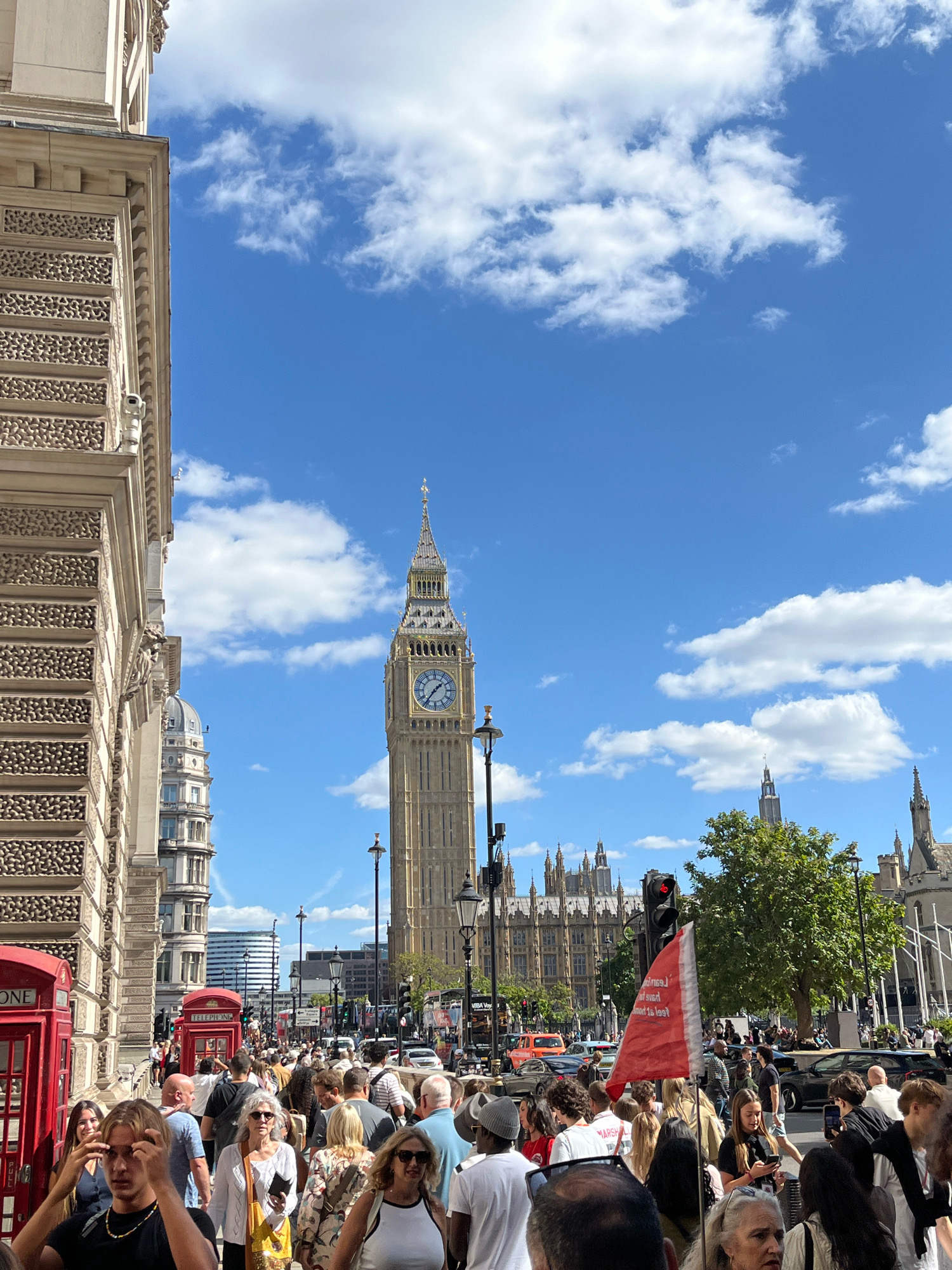 The image depicts a street scene in Westminster, London, United Kingdom, with the iconic Elizabeth Tower (commonly known as Big Ben) prominently featured in the background. A large crowd of people is walking along the street, suggesting a busy tourist area or event.
In the foreground, a young man with blonde hair is looking towards the camera, touching his hair. To his left are red telephone booths. Several other individuals are visible in the crowd, some looking at their phones, others engaged in conversation. A red flag with white text is held up by someone in the crowd.
The environment is urban, with ornate architecture of buildings lining the street. The sky is blue with scattered white clouds, indicating fair weather. Vehicles, including buses and cars, are visible on the street, suggesting active city life. The overall impression is one of a bustling and popular tourist destination in London.