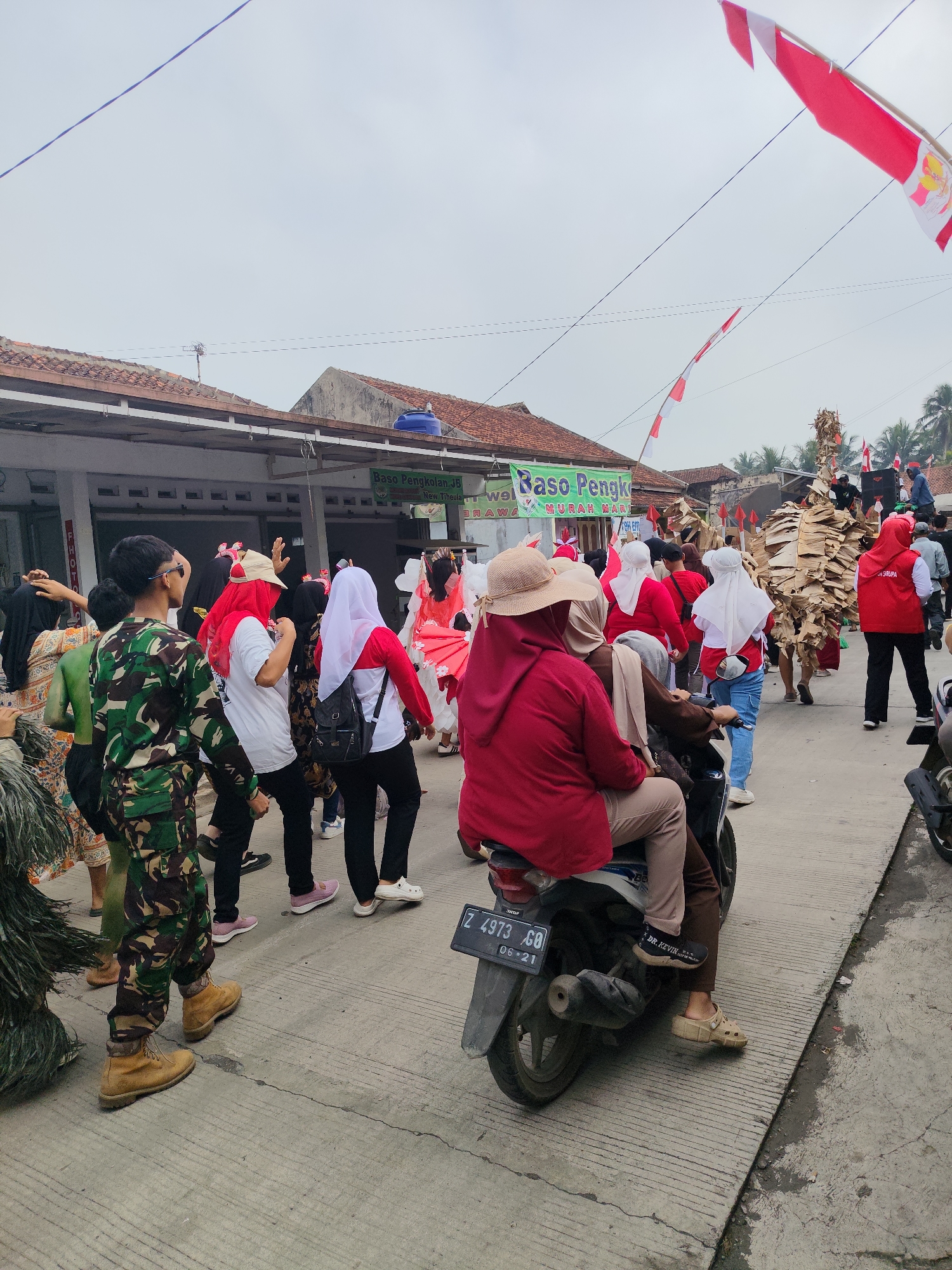 The image depicts a street scene in Sukaratu, Indonesia, during a parade or procession. The foreground features a motorcycle with two people, a woman wearing a red hijab and riding on the back, and a person in a tan hat and red top driving. The motorcycle has a license plate that reads "Z 4973 GO 06-21". To the left of the motorcycle, a person in a camouflage uniform walks forward, wearing brown boots. Behind them, several people are walking, some in red and white attire, and others in colorful outfits. In the background, buildings line the street, and red and white flags are visible, along with palm trees. One building has a green sign that reads "Baso Pengkolan JB" and "MURAH MERIAH". Another person in the background is wearing a costume made of cardboard. The overall atmosphere suggests a community event or celebration.