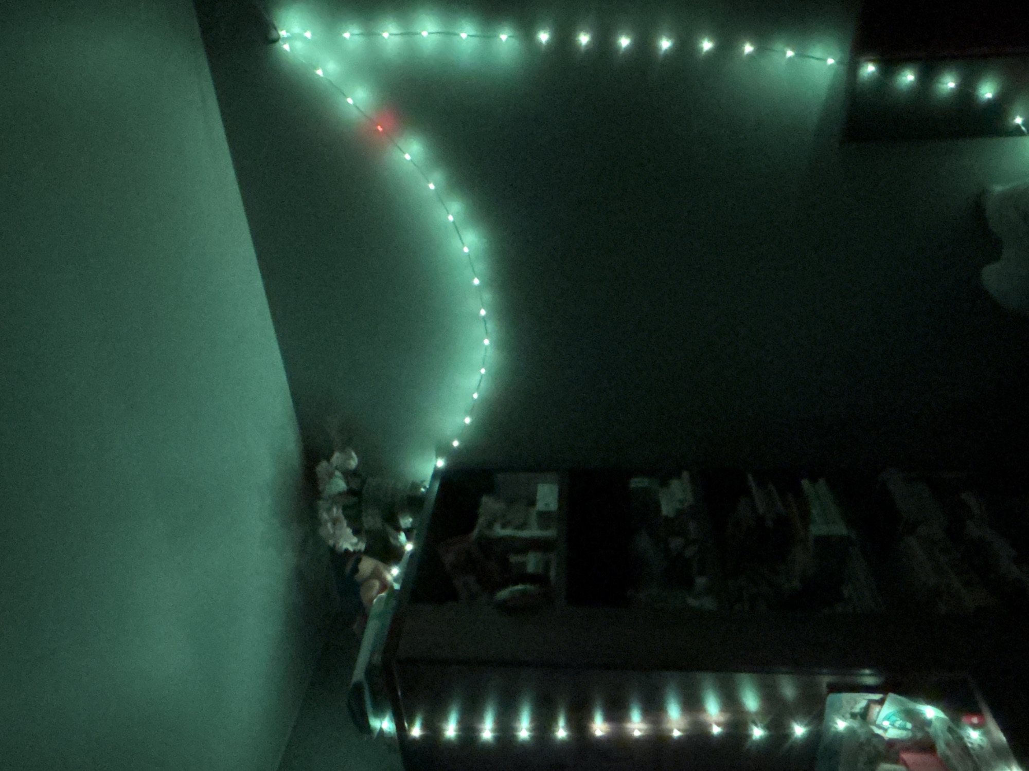 The image depicts an indoor scene, likely a bedroom, illuminated by string lights. The dominant color is a deep green, cast by the lights themselves and possibly the surrounding environment. On the left side of the frame, a dark bookshelf is visible, its shelves partially obscured by darkness and the glow of vertical strings of white and teal lights. On top of the bookshelf, a bouquet of light-colored flowers is arranged in a vase, with some objects beneath the flowers. A string of green lights trails across the upper wall and then down the right-hand wall, with one red light interspersed within the green ones. The overall impression is one of a dimly lit, possibly decorated, room. There are no individuals present in the image. The location of Novaya Usman', Russia, is not discernible from the visual information provided in the image.