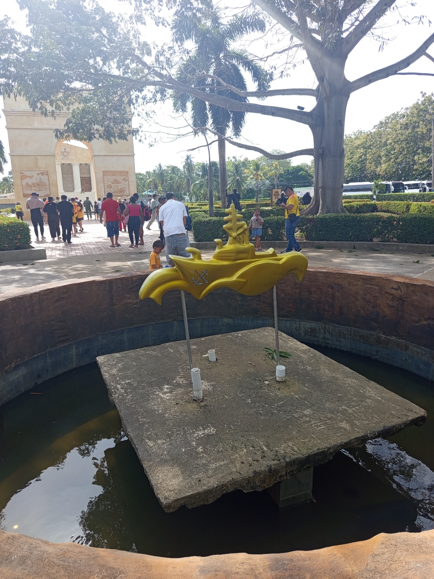 In Coveñas, Colombia, a group of people are gathered near a large stone archway. In the foreground, a yellow, genie lamp-shaped sculpture is positioned on a concrete platform within a water feature. The sculpture has anchor motifs. The environment is sunny with lush green trees and foliage surrounding the area. A large, old tree with thick branches dominates the background. Several buses are parked in the distance.