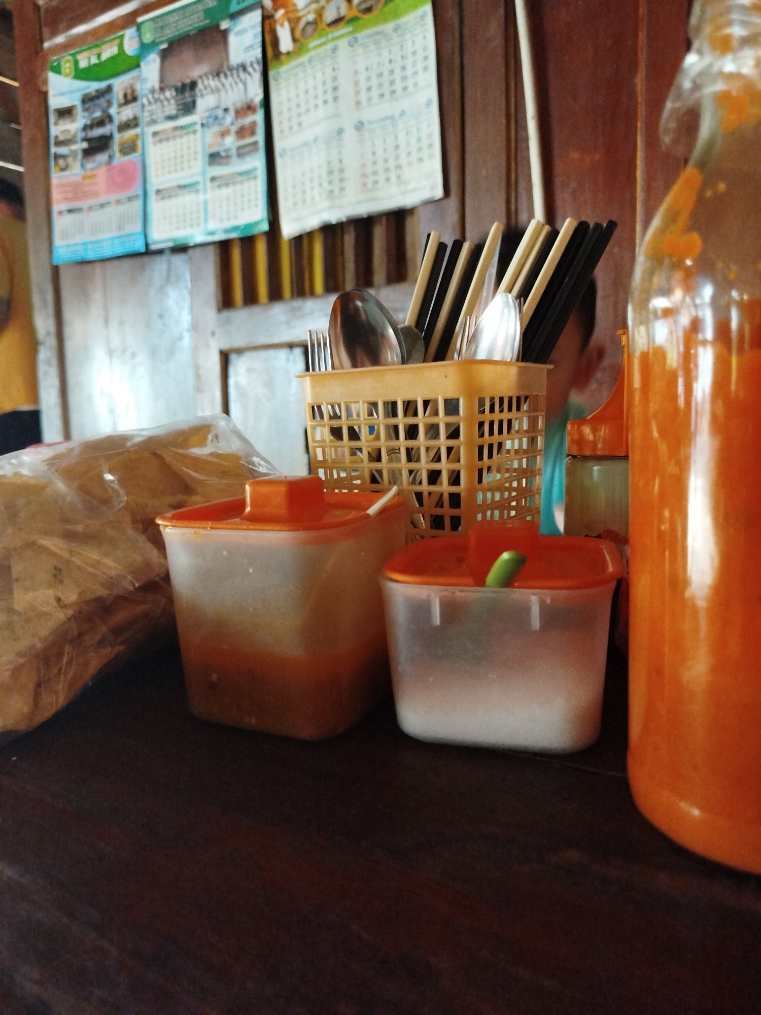 The image depicts a close-up shot of a table at what appears to be a food establishment in Pojok, Indonesia. The foreground is dominated by a dark wooden table surface. On the table, there are several objects: to the left, a clear plastic bag filled with what looks like snacks or food items is partially visible. Next to it, two clear plastic containers with orange lids are present. The container on the left contains a reddish-orange substance at the bottom and appears to be filled with liquid or sauce. The container on the right is filled with a white granular substance, likely salt or sugar, and has a small green object, possibly a chili or garnish, protruding from its lid. In the middle ground, a yellow plastic basket holds an assortment of chopsticks and spoons. Behind the basket, a large, clear glass bottle, mostly filled with an orange liquid, is prominently featured on the right side of the image. In the background, a wooden wall is visible, adorned with several calendars and posters, suggesting a rustic or traditional setting. The overall scene is casual and indicative of a mealtime setting at a local eatery.