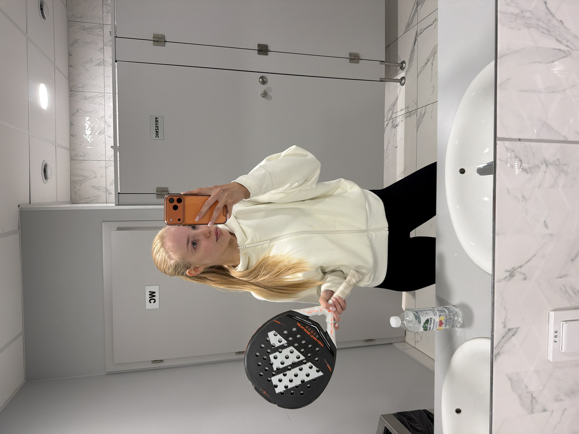 A young woman is taking a selfie in a public restroom, likely in the city of Unknown, Unknown. She is holding an orange smartphone in her right hand. Her left hand is holding a black and white padel racket. She is wearing a white zip-up hoodie and black leggings. The background shows restroom stalls with marble-patterned walls, white sinks, and a trash bin. A bottle of water is sitting on the sink counter.