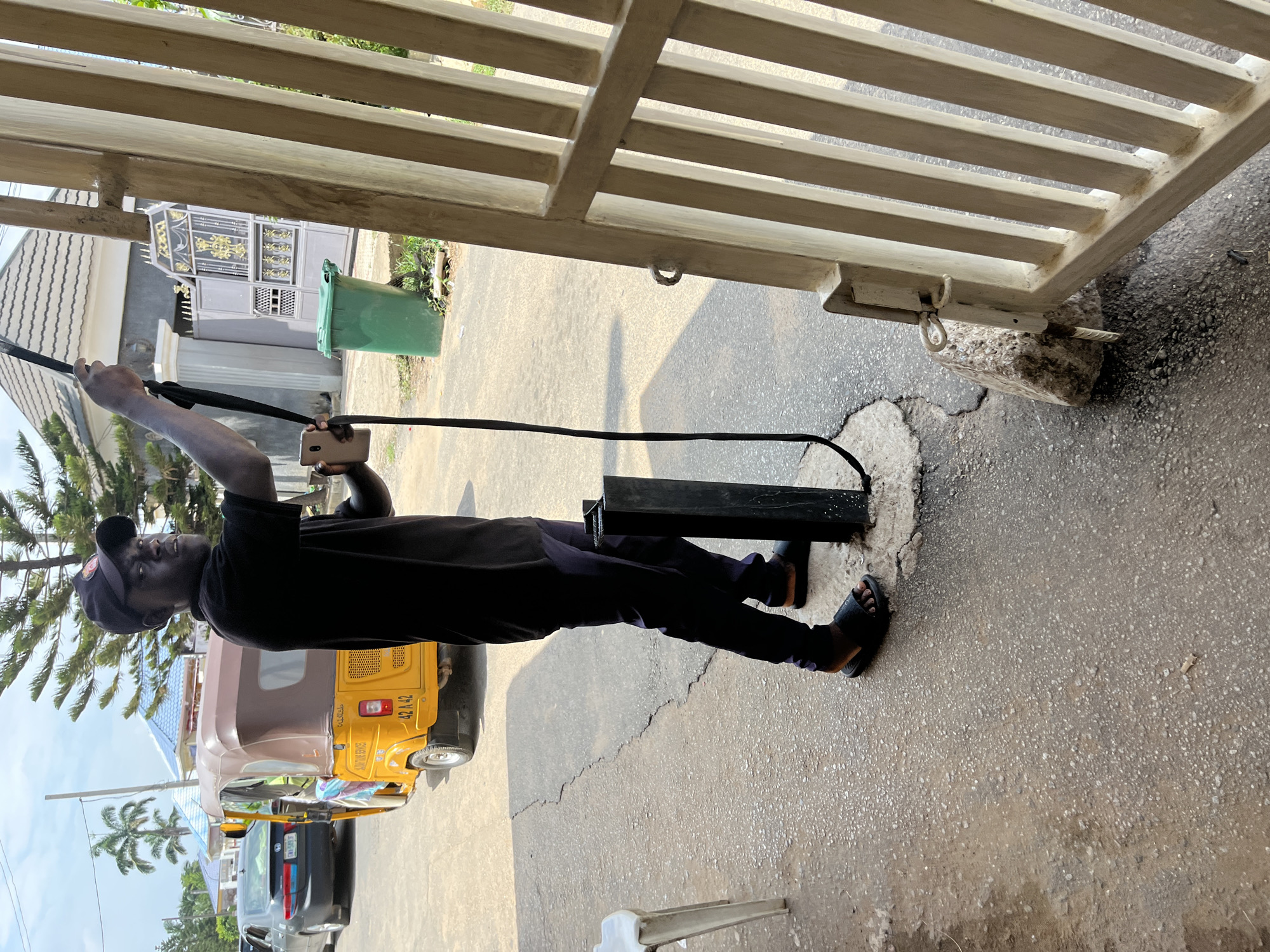 The image depicts an outdoor street scene in what appears to be a residential area of Abuja, Nigeria, during daylight hours under a bright, partly cloudy sky.
In the foreground, a Black male, approximately 20-30 years old, stands facing slightly right, looking towards the camera. He wears a black short-sleeved polo shirt, dark trousers, and black slide sandals. A black baseball cap with a red logo is on his head. His left hand holds a light-colored smartphone, and his right hand holds a long, black strap or cable that extends downwards to a black metal post anchored in a concrete base on the ground.
To the immediate right, a white, vertical-slatted metal gate is partially open, propped by a light-colored concrete block. A metal ring is affixed to the inner frame of the gate.
Behind the man, on an asphalt road, a yellow three-wheeled motorized rickshaw, commonly known as a Keke Napep, is visible. It has a brown canopy, the text "AUN SERVICE" partially visible on its side, and "42 A 42" printed on its rear. Further down the road, a dark-colored sedan, identified as a Honda Accord, is parked, displaying a Nigerian license plate "DW 525 EJ."
In the background, beyond the road, residential structures are visible. One building features a grey tiled roof and an elaborate grey and gold metal gate. Another building further back has a light blue roof. A green wheeled trash bin is positioned on the roadside near the ornate gate. Several palm trees and other foliage are present throughout the background. The ground is a paved surface, exhibiting cracks and wear. A partial white plastic chair is visible on the far left edge of the image.
The scene represents a typical urban or suburban environment in Abuja, Nigeria, characterized by local transportation (Keke Napep) and residential architecture. The precise location within Abuja cannot be determined from the provided visual information alone.