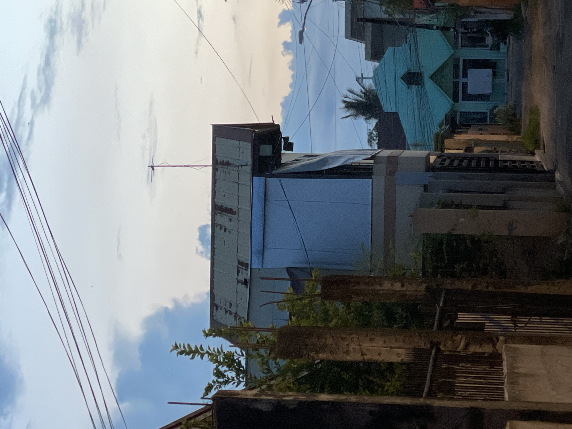 This is a photograph taken in Davao City, Philippines, likely in a residential or mixed-use area, featuring a street scene at dusk. Dominating the foreground are the edges of buildings and fences, with a partial view of a concrete wall and a metal gate on the left. The central and right portions of the image reveal a street receding into the distance.
Further down the street, a bright turquoise building with a steeple and cross, resembling a small church or chapel, is visible. It has a distinct architectural style with gabled roofs and multiple windows. To the left of this building, another structure with a corrugated metal roof and a light blue exterior is partially seen, featuring what appears to be a tarp or large sheet covering part of its facade. Electrical wires crisscross the sky, and an antenna is mounted on top of one of the buildings.
The sky is filled with a mix of blue and scattered clouds, with hints of sunset colors at the horizon. The overall impression is that of a typical neighborhood in the Philippines during the transition from day to night, characterized by its residential architecture and overhead utility lines. There are no people visible in the frame.