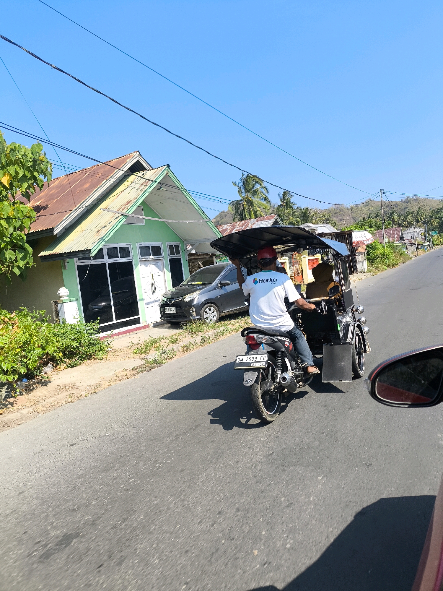 A motorized tricycle, or "becak motor," is traveling on a paved road in Tilamuta, Indonesia, on a sunny day. The driver, wearing a red helmet and a white t-shirt with a "Harka PAINT" logo, is steering the vehicle. A passenger is seated behind the driver, visible through the fabric canopy of the tricycle. To the left of the road, a small, light green house with a corrugated metal roof is situated. A dark gray car is parked in front of the house. The environment is rural, with vegetation lining the roadside and rolling hills in the background. Overhead power lines crisscross the bright blue sky. The scene is captured from the perspective of another vehicle traveling behind the tricycle.