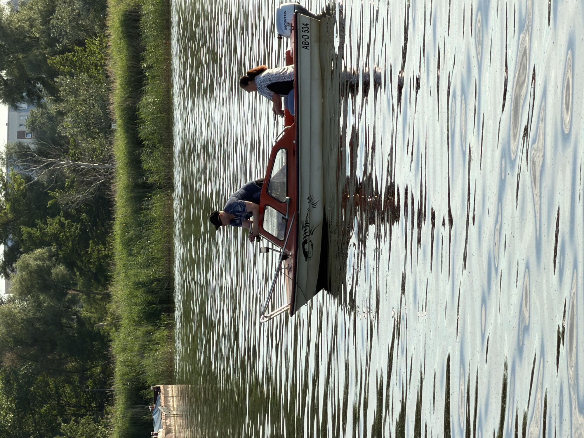 The image depicts two people in a small motorboat on a body of water, likely a lake or river, near a grassy bank with trees. The boat is white with red trim and has a Honda outboard motor. Visible text on the boat reads "AB-D 534". The person in the back, a woman with dark hair, is leaning forward, possibly operating the boat or observing something. The person in the front, a man, is seated and looking down. The water is rippled, suggesting a gentle breeze. The sun appears to be out, casting reflections on the water and indicating a daytime setting. The lush green of the bank and trees suggests a warm season, likely spring or summer. In the background, the faint outline of buildings can be seen, hinting at a proximity to a town or settlement. The overall scene is tranquil, suggesting a leisurely outing. The location is identified as Ribnitz-Damgarten, Germany.