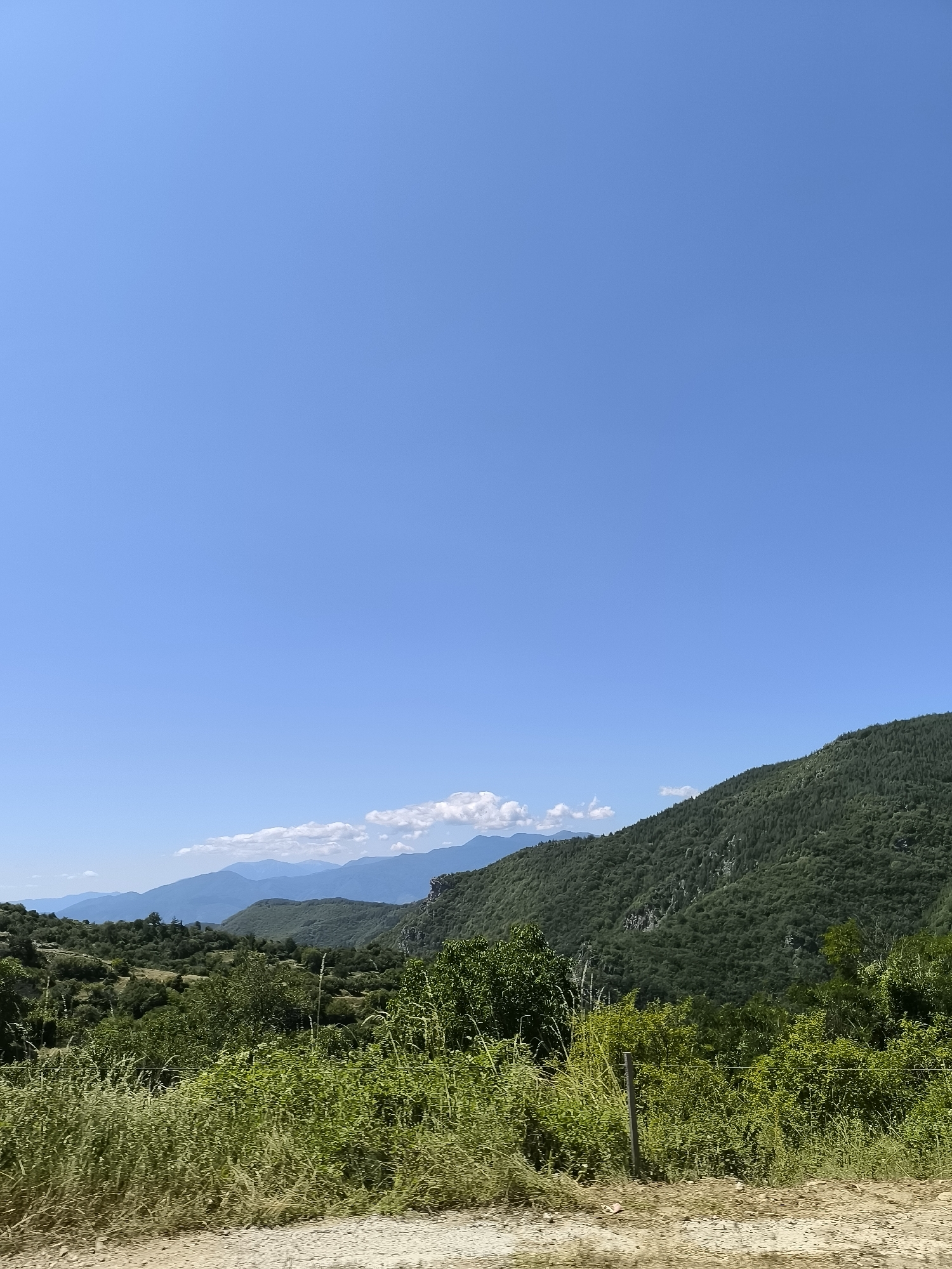 The image depicts a vast, clear blue sky dominating the upper half of the frame. Below the sky, a lush, green mountainous landscape unfolds. Rolling hills and slopes are covered in dense vegetation, with trees and shrubs creating a textured surface. In the foreground, a dirt road or path is visible, bordered by tall, dry grass and smaller green plants. A simple wooden post with a thin wire fence runs horizontally across the lower part of the scene, separating the road from the denser foliage. In the distance, layers of mountains recede, with softer blue hues indicating their distance. Wispy white clouds are scattered among the distant peaks. The scene is clearly an outdoor, natural environment, likely a rural or mountainous area, consistent with the location in Leshten, Bulgaria, known for its picturesque natural beauty and traditional architecture. There are no people or animals visible in the image.