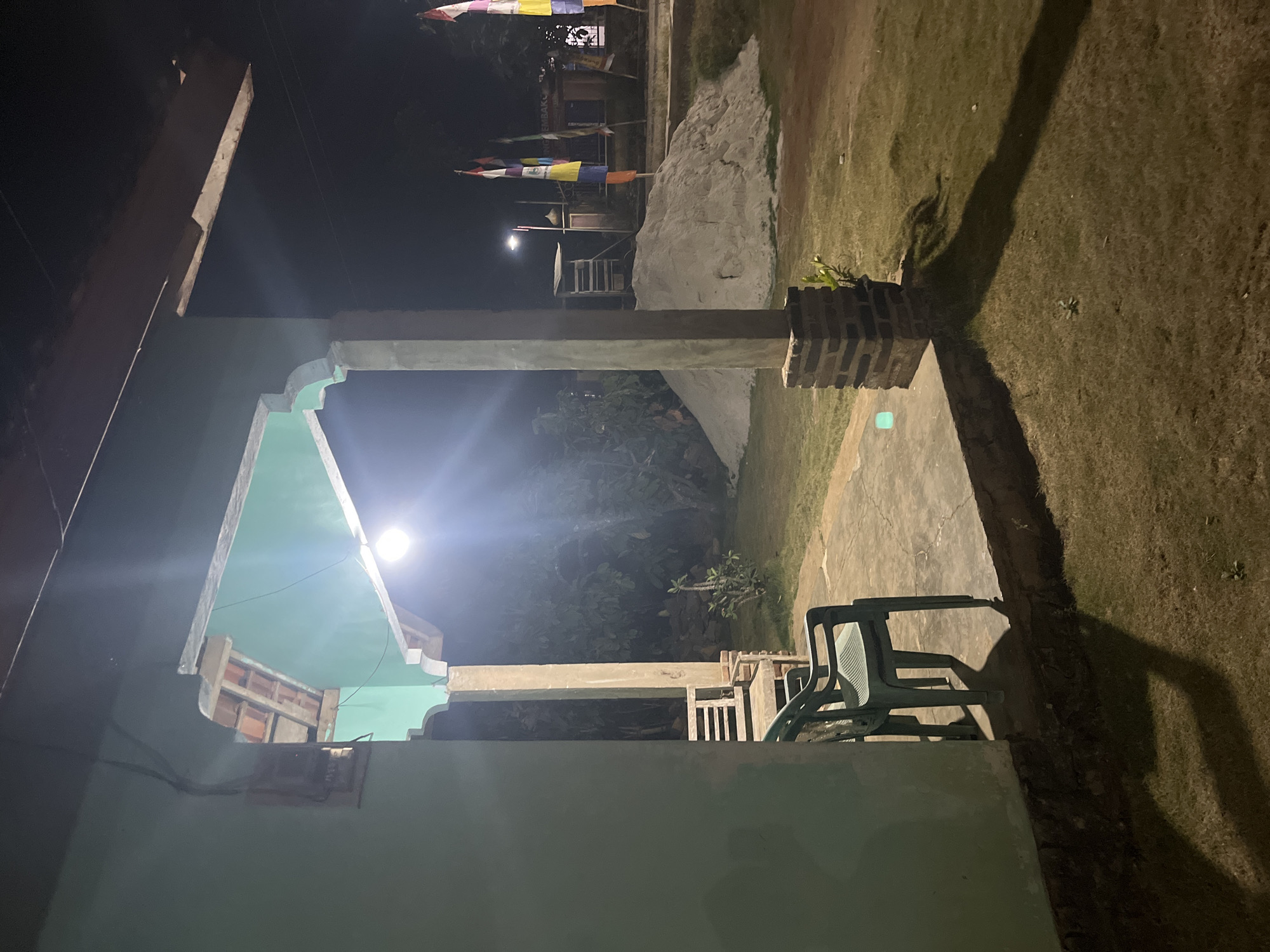The image captures an outdoor scene at night, likely in Kebumen, Indonesia, characterized by a tranquil, dimly lit atmosphere. The foreground is dominated by a light green wall on the left, suggesting a building or structure. Adjacent to this wall is a concrete porch area, upon which rests a dark green plastic chair and the partial view of another seating object, possibly a bench. The porch itself is elevated, with a lower concrete step. A thick concrete pillar, constructed with brick at its base, stands to the right of the porch, supporting an overhead structure with decorative arches.
Beyond the porch, a grassy yard extends into the background, illuminated by a bright overhead light fixture casting stark shadows. In the middle ground, a large mound of light-colored sand is visible. Behind the sand mound, a wooden ladder-like structure and a building with colorful flags are discernible. The surrounding environment appears to be filled with dense foliage and trees, adding to the evening ambiance. The overall impression is one of a rural or semi-rural setting, possibly a guesthouse or residential area, at rest during the night.