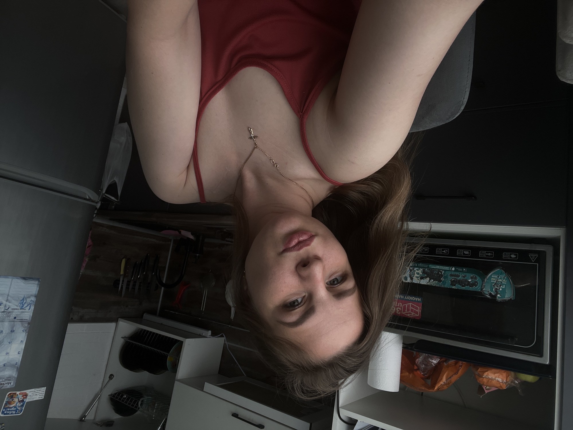 A woman with light brown hair and fair skin, wearing a red tank top and a delicate necklace, is positioned centrally in the image. She is facing the camera with a neutral expression. The background reveals a kitchen setting. A refrigerator is partially visible to the left, and a rack containing various kitchen utensils is mounted on the wall. Lower kitchen cabinets and a microwave oven are also visible. A roll of toilet paper is positioned between the microwave and a countertop. The image appears to be taken indoors. There is no explicit information to confirm the location as Izhevsk, Russia in this image.