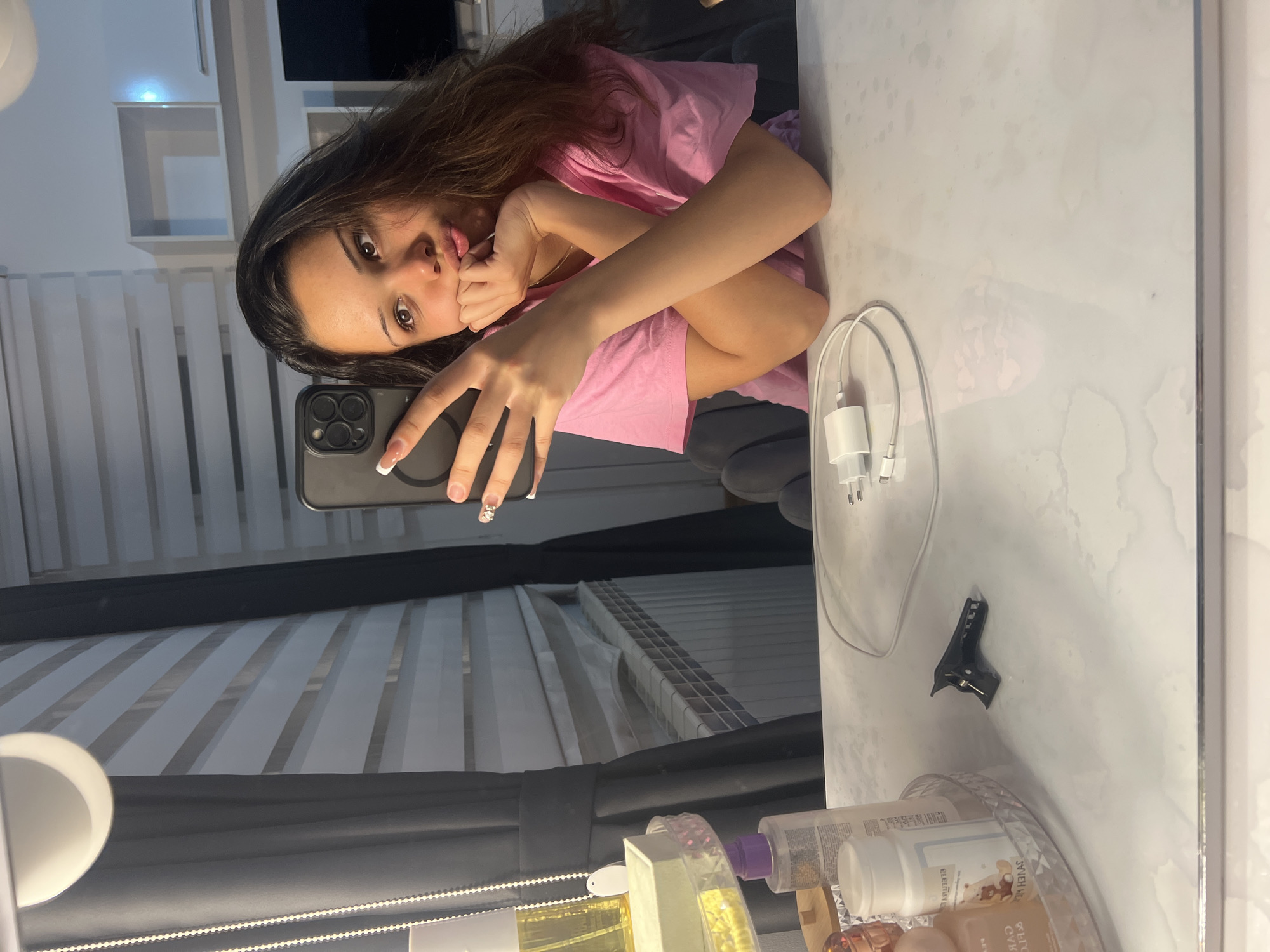 A young woman is captured in a selfie taken in a mirror or highly reflective surface, giving the impression of an inverted shot due to the phone's position. She has long, dark brown hair and is wearing a pink t-shirt. Her expression is playful, with her tongue slightly out, and her right hand is near her mouth. She holds a smartphone with a black case and visible camera lenses in her left hand, which has well-manicured nails featuring a French tip design and some embellishment on one finger.
The setting appears to be an indoor room, possibly a bedroom or dressing area. In the foreground, on the white, glossy surface where the reflection is seen, several objects are visible. These include white charging cables and a power adapter, a black hair clip, and a collection of bottles and containers, likely beauty products, arranged on a clear, faceted tray. Text on some bottles is partially legible; one appears to say "ESSENCES" or "ELISIR", and another with brown liquid has Cyrillic script, possibly indicating "УВЛАЖНЯЮЩИЙ".
The background behind the woman reveals blinds with horizontal slats and some empty white shelves or cubbies. A small, circular white light fixture is also partially visible on the left side of the reflection. The overall impression is casual and personal, with the bright lighting suggesting an interior space.