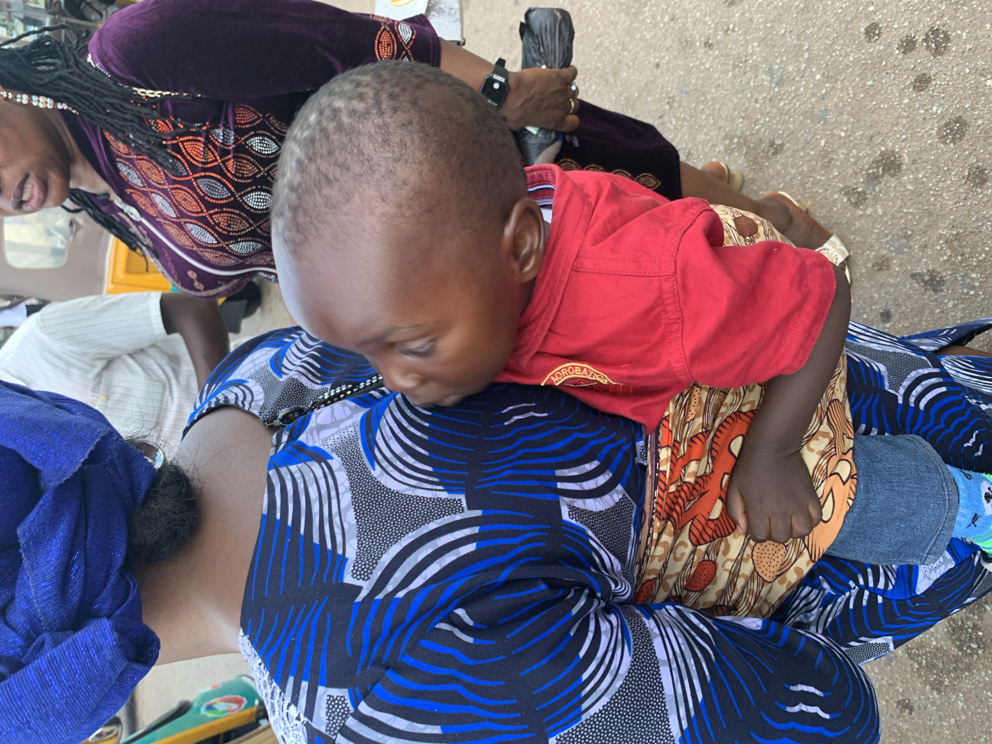 A close-up, medium shot shows a baby being carried on the back of a woman, as seen from the perspective of the viewer looking slightly down. The baby, wearing a red shirt, faces towards the right and appears to be nuzzling into the carrier's shoulder. The carrier, a woman with a blue headwrap and a blue, black, and white patterned dress, is seen from the back, with her head turned to the right, her neck and shoulder visible. A second woman, wearing a purple velour jacket with an embroidered pattern and large earrings, stands to the right of the carrier, her face partially visible and her mouth open as if speaking. She is holding a black plastic bag in her right hand. Both women are wearing jewelry. The environment appears to be an outdoor, possibly urban setting, with a paved surface underfoot and hints of what might be a street or market, including a blurred yellow vehicle in the background. The lighting is natural and the image is sharp, capturing the textures of the fabrics and the skin tones of the individuals. The scene conveys a sense of everyday life and maternal care.