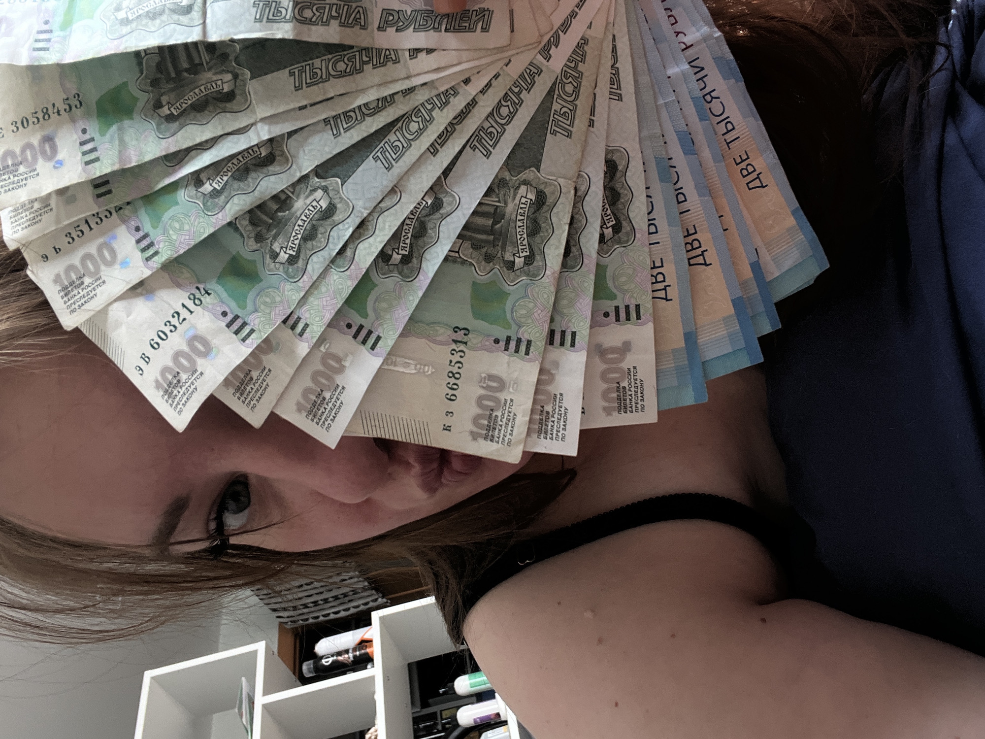 A woman is holding a fan of Russian banknotes in front of her face. The bills are a mix of 1000 ruble notes (with "ТЫСЯЧА" printed on them and a depiction of Yaroslavl) and 2000 ruble notes (with "ДВЕ ТЫСЯЧИ" printed on them). The woman's face and shoulder are visible. There is a white shelving unit visible in the background. The location is not mentioned in the image and cannot be identified.