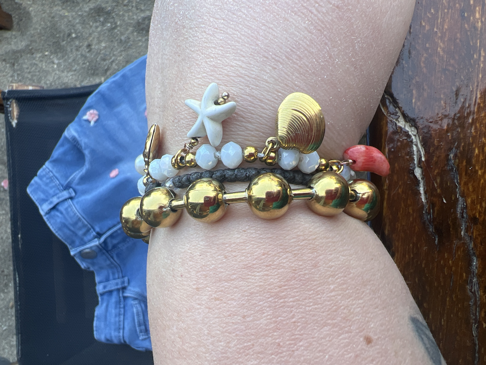 The image shows a close-up of a person's arm adorned with multiple bracelets. The bracelets are a mix of gold-tone beads, white faceted beads, and dark gray cord, with various charms including a white starfish, a gold-tone seashell, and a red coral piece shaped like a bean. The arm appears to be resting on a dark wooden surface. In the background, a portion of blue denim shorts is visible. The scene is outdoors, with a slightly blurred background suggesting natural lighting and an outdoor environment. No specific location in Eretria Municipal Unit, Greece is identifiable from the image.