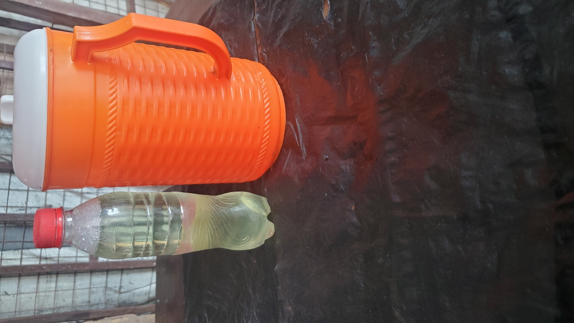 In Jalingo, Nigeria, an orange plastic pitcher with a white lid and a handle sits next to a clear plastic bottle filled with a light yellow liquid. Both items are placed on a dark, textured surface that appears to be a tablecloth or tarp. In the background, a blurred metal grate or fencing is visible. The lighting suggests an outdoor or dimly lit indoor environment.