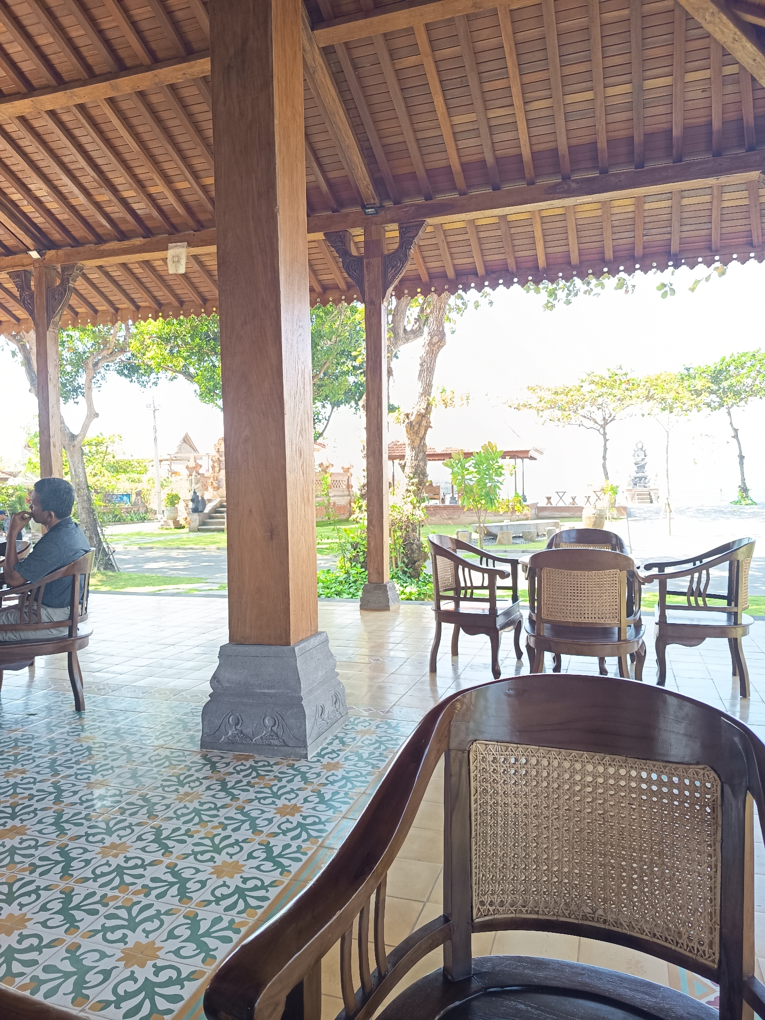 The image depicts an outdoor seating area, likely a cafe or restaurant, with traditional Indonesian architecture. A man in a gray shirt is seated at a wooden chair, looking towards the left. In the background, there are more wooden chairs and tables, a garden with trees, and a glimpse of what appears to be a traditional Balinese temple structure. The floor is tiled with a decorative pattern of green and yellow motifs. The overall scene suggests a serene and culturally rich environment in Denpasar, Indonesia.