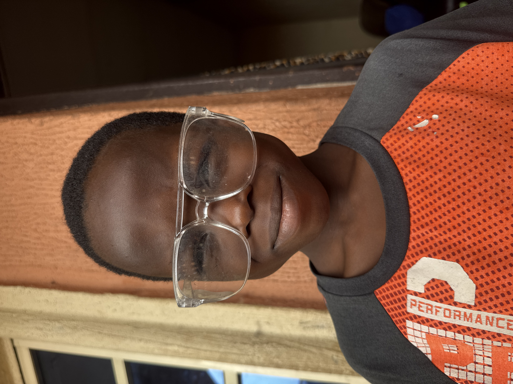 A person with dark skin is wearing clear-framed glasses and an orange and gray shirt with the word "PERFORMANCE" printed on it. The person's eyes are closed. The background includes a textured, peach-colored wall and a window. The image is taken indoors. There is no specific information to indicate the location in the city of Onitsha, Nigeria.